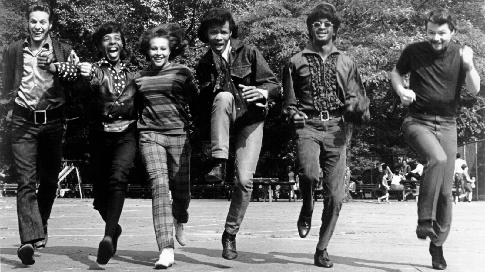 band 桌布 壁紙 — 六個穿著1970年代時尚的人，在陽光普照的公園廣場上充滿活力地跳躍，背景有樹木和長椅。
