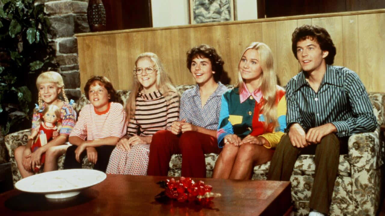 show 桌布 壁紙 — 六個穿著1970年代服裝的人一起坐在一個舒適客廳的花卉沙發上，客廳有石牆和木鑲板。