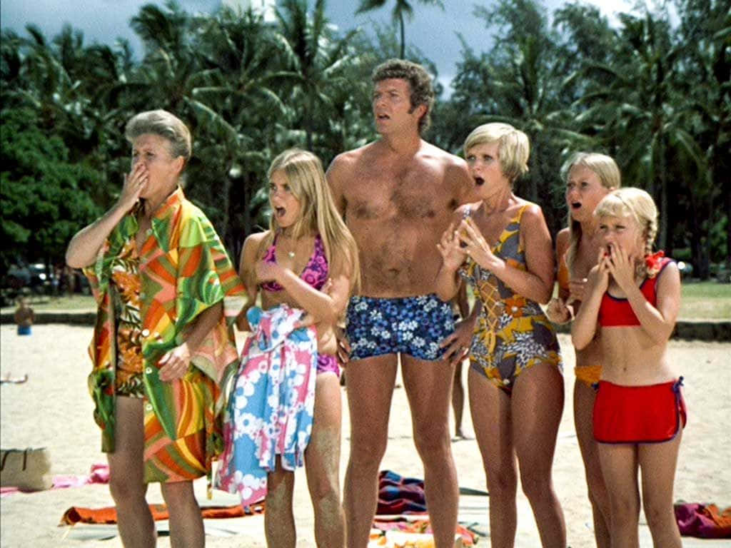 show 桌布 壁紙 — 六個穿著1970年代沙灘裝的人在沙灘上震驚地倒抽一口氣，後面有棕櫚樹。
