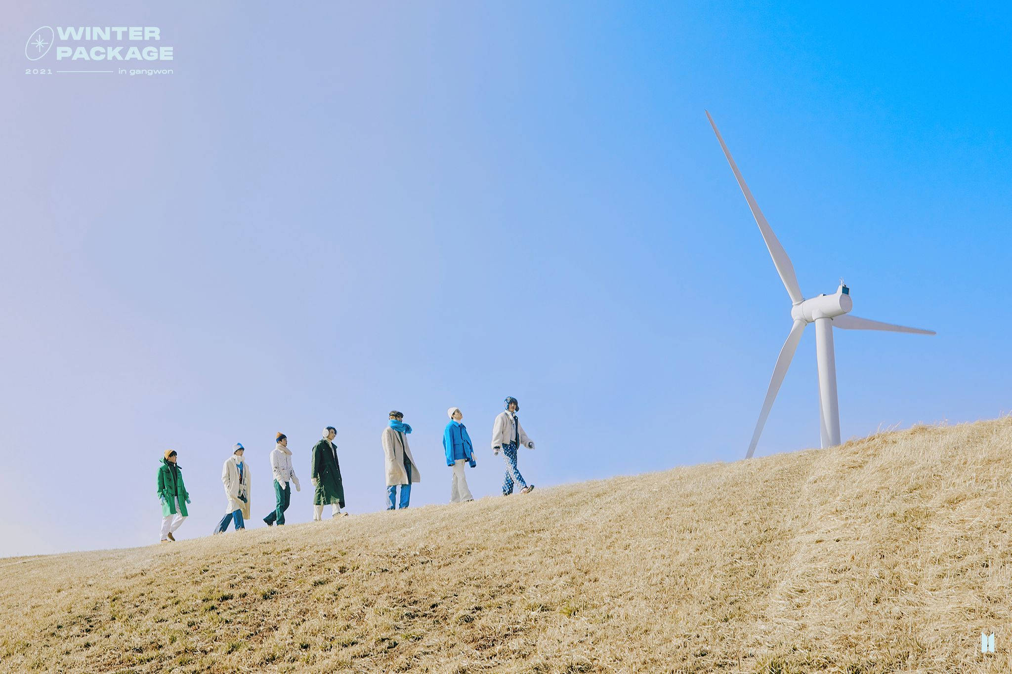 BTS 2021 桌布 壁紙 — 六個穿著冬季外套的人在一片乾燥的草地上走過，背景是晴朗的藍天，附近有一座高大的白色風力發電機。