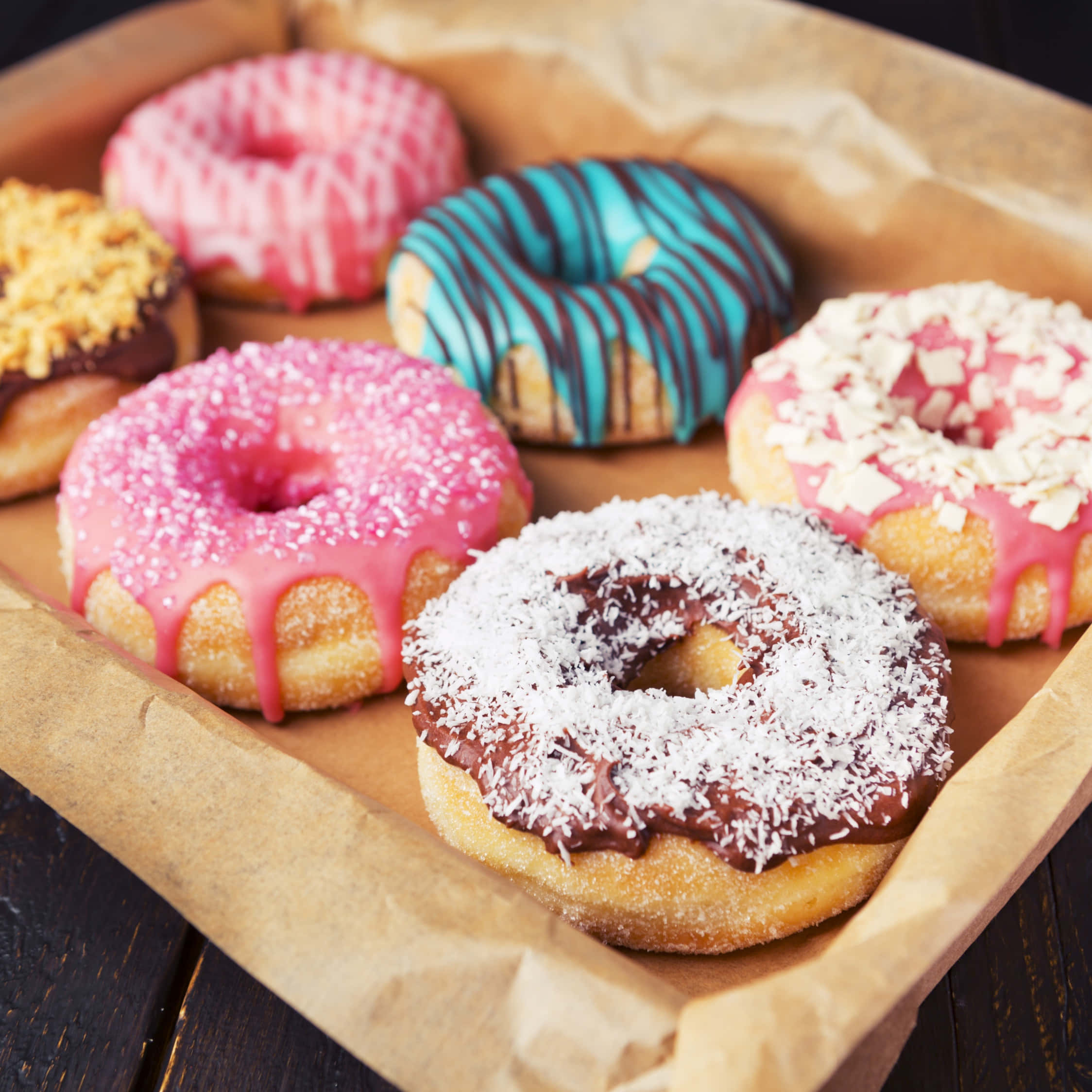 Cute Donut 桌布 壁紙 — 六個色彩鮮豔的釉面甜甜圈，上面有粉紅色、藍色、巧克力和椰子配料，放在一個鋪有棕色紙的托盤上，置於深色木質表面。