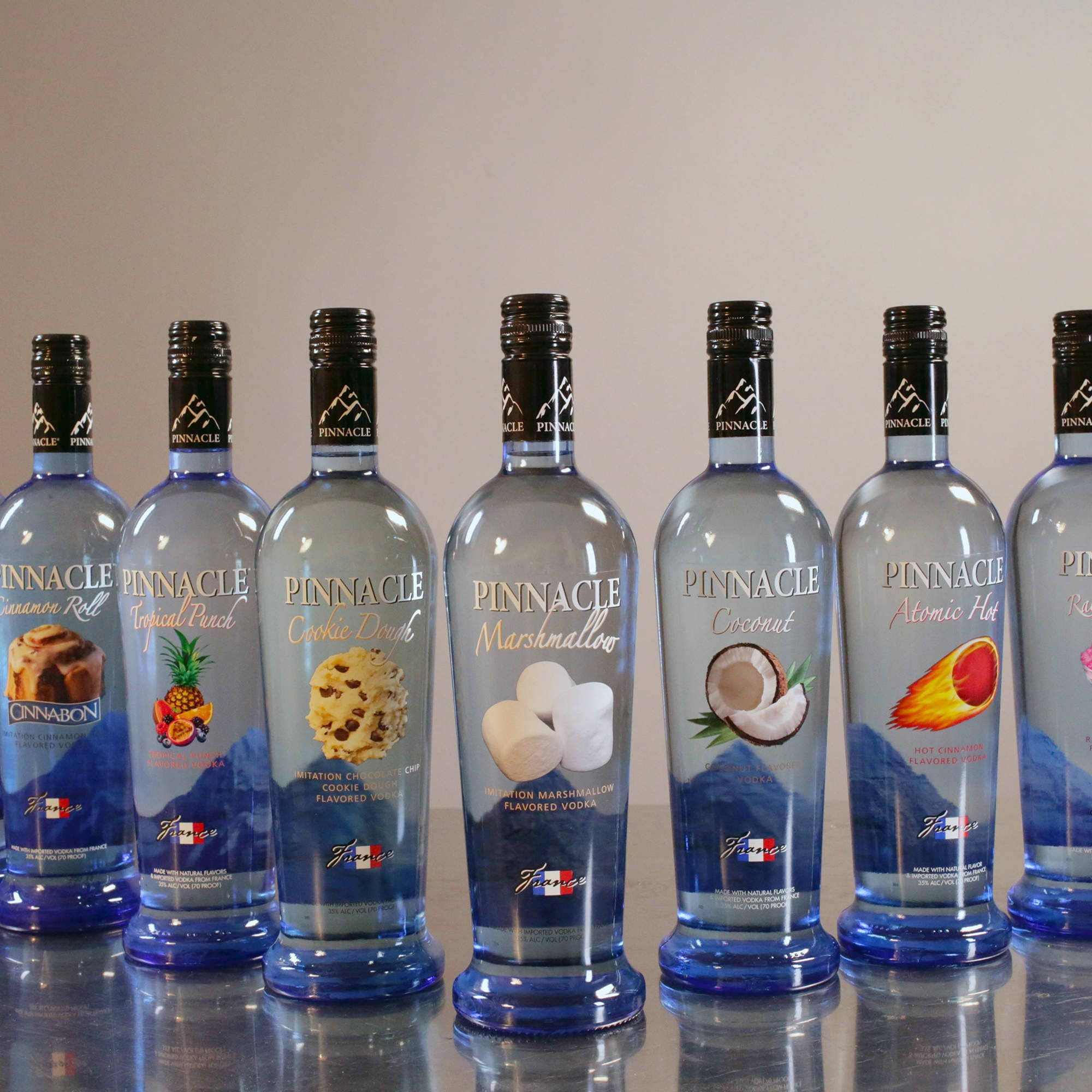 bottle 桌布 壁紙 — 六個藍色 Pinnacle 伏特加酒瓶帶有口味標籤，排成一排放在反射表面上，背景為素牆。