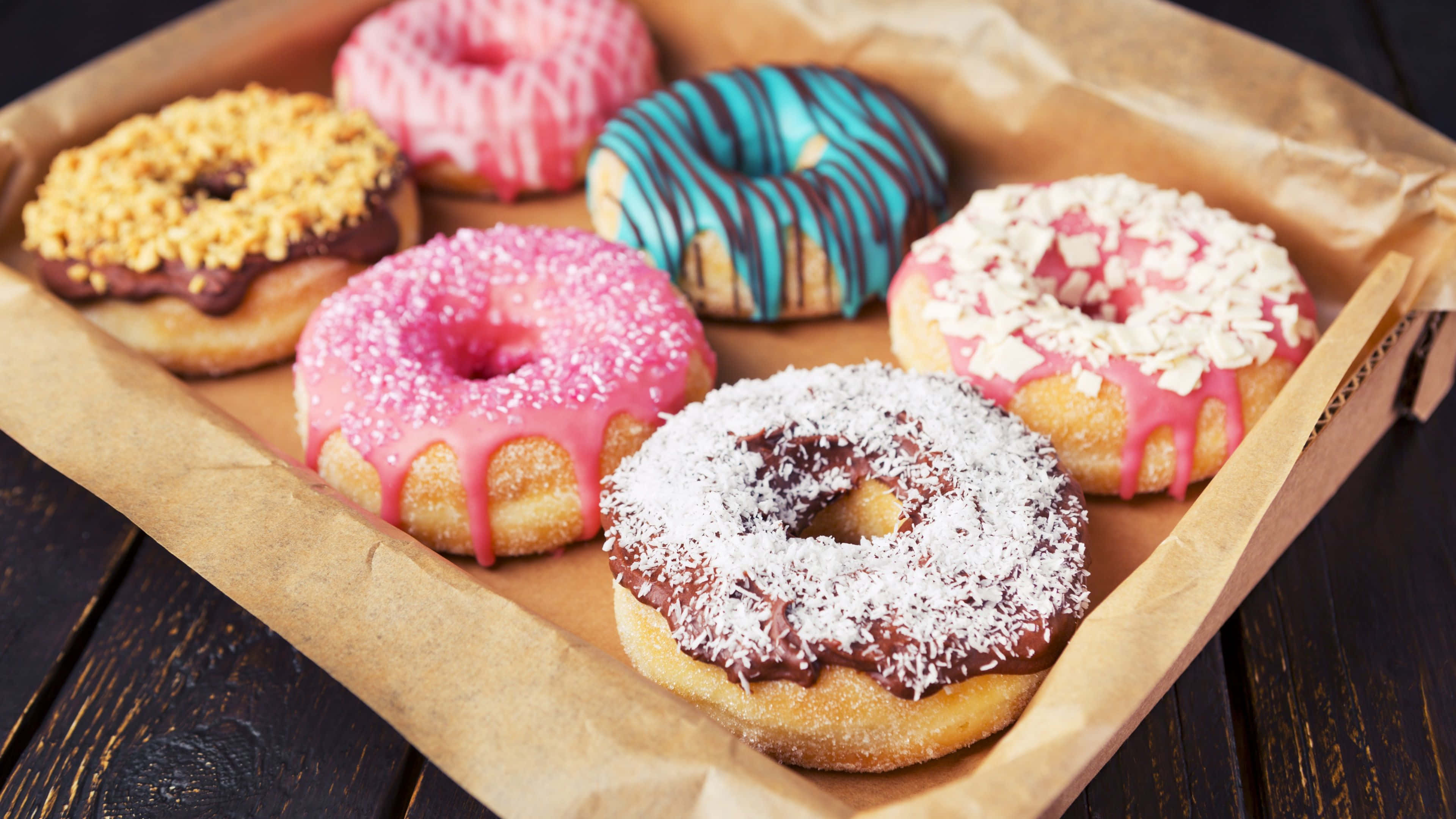 Dunkin Donuts 桌布 壁紙 — 六個裝飾鮮豔的甜甜圈，有粉紅色、藍色和巧克力糖霜，放在深色木桌上的棕色紙托盤上。