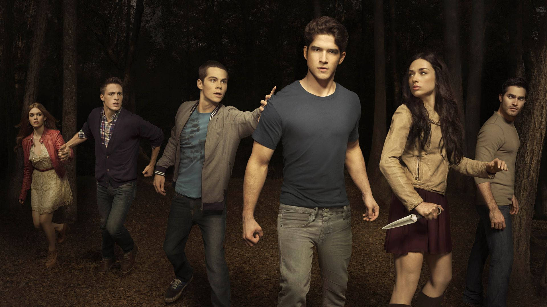 scott mccall 桌布 壁紙 — 六名年輕成年人在黑暗的森林中，緊張警覺，手持武器，彷彿在逃離或面對危險。