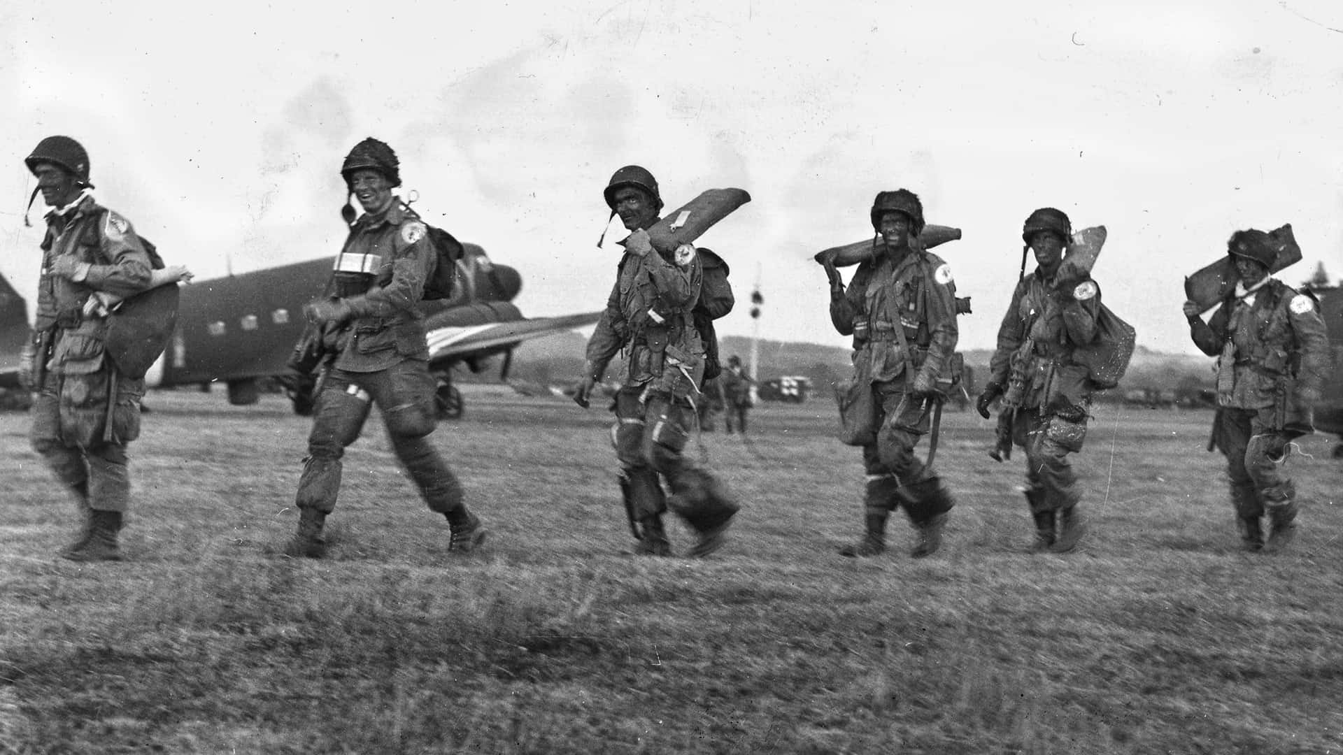 Ww2 桌布 壁紙 — 六名微笑的傘兵在草地上行軍，背景有軍用運輸機的黑白照片。