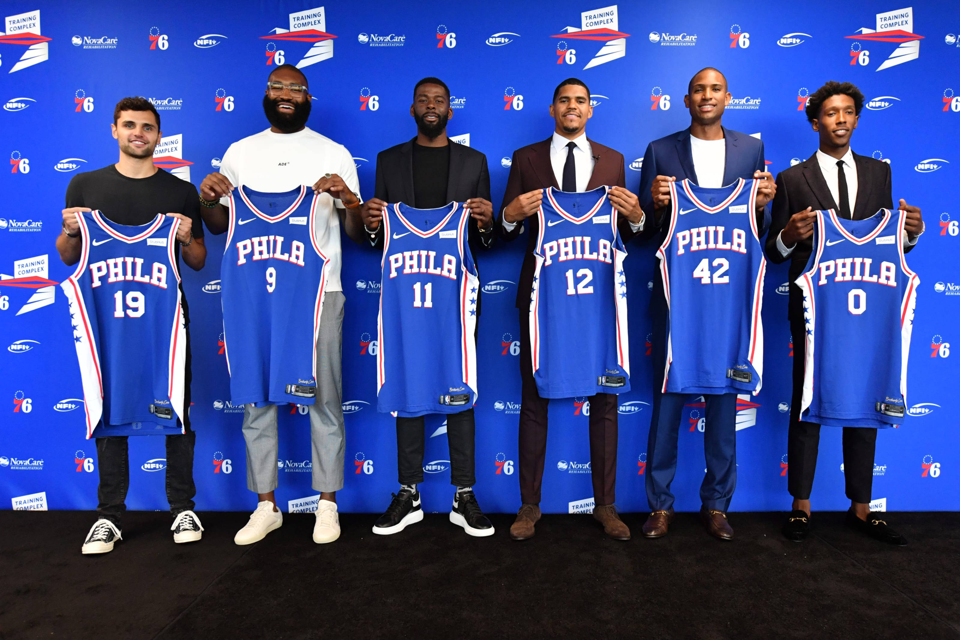 76ers 桌布 壁紙 — 六名籃球運動員在藍色品牌牆前擺姿勢，每人舉起一件帶有不同號碼的藍色費城76人球衣。