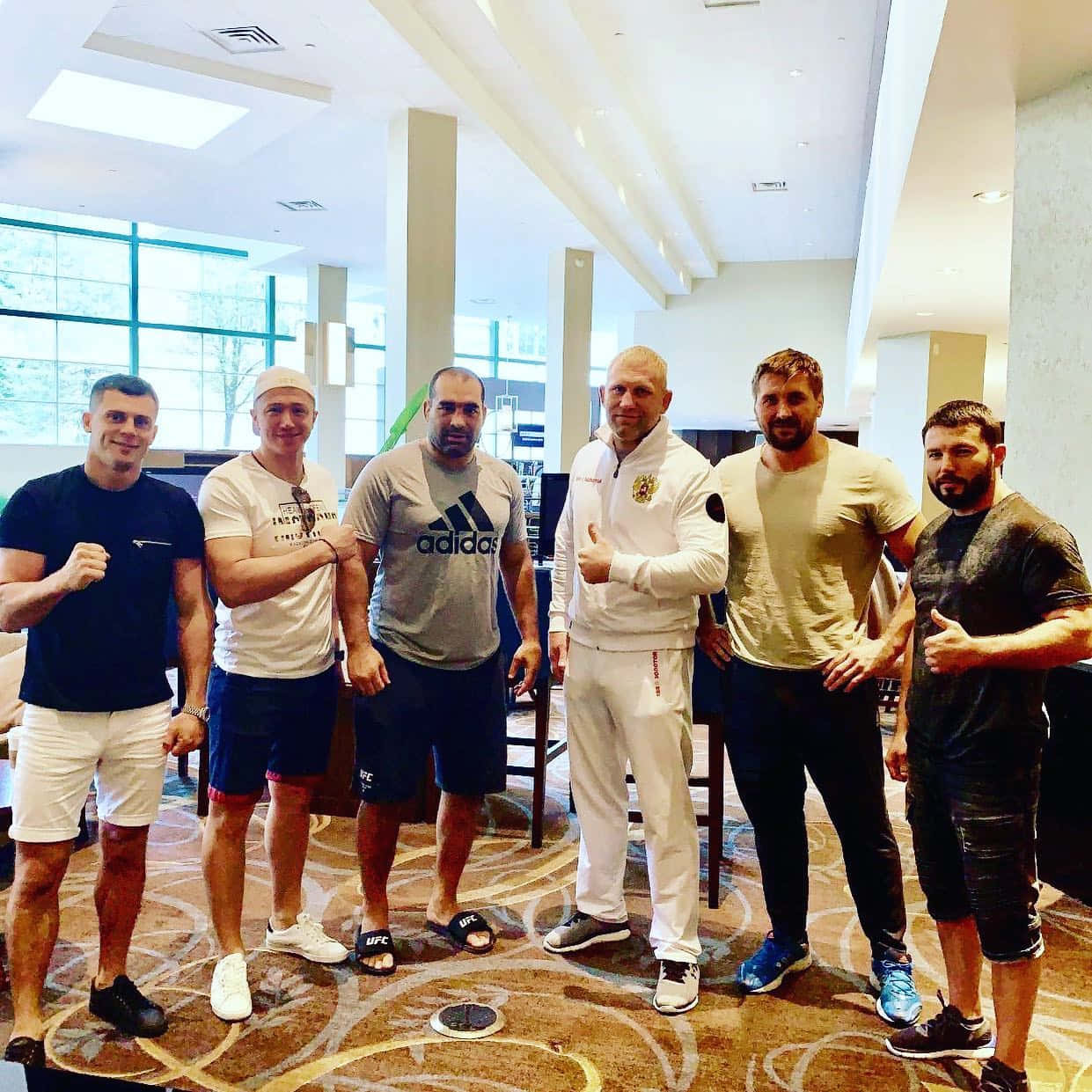 UFC 桌布 壁紙 — 六名肌肉發達的男子穿著運動服，在設有大窗戶和圖案地毯的現代飯店大廳自信地擺姿勢。