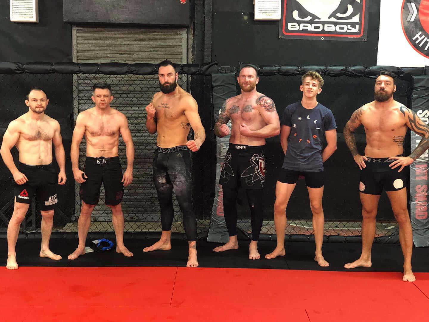 Fitness 桌布 壁紙 — 六名肌肉發達的男子身穿格鬥裝備，在鋪有紅色軟墊的MMA鐵籠內自信地擺姿勢，身後牆上印有「BADBOY」品牌標誌。