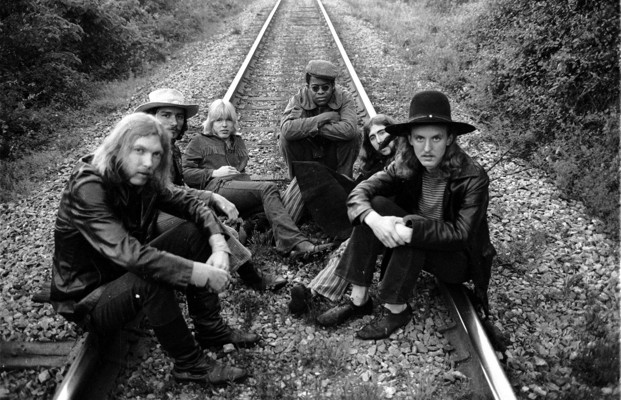 band 桌布 壁紙 — 六名身穿1970年代服裝的男子坐在和蹲在鐵路軌道上，兩旁是茂密的樹葉，一張清晰的黑白照片。