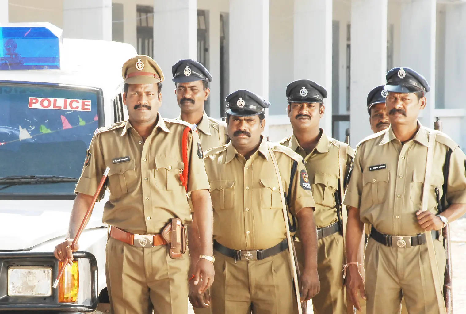 Indian 桌布 壁紙 — 六名身穿卡其色制服、表情嚴肅的警察自信地站在一輛帶有藍色燈光的白色警車前。