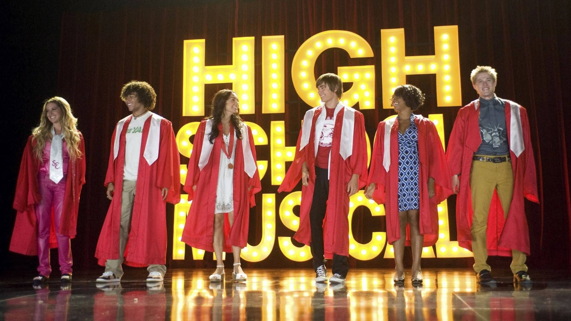 high 桌布 壁紙 — 六名身穿紅色學士帽和畢業袍的畢業生，在發光的「HIGH SCHOOL MUSICAL」標誌前微笑站在舞台上。