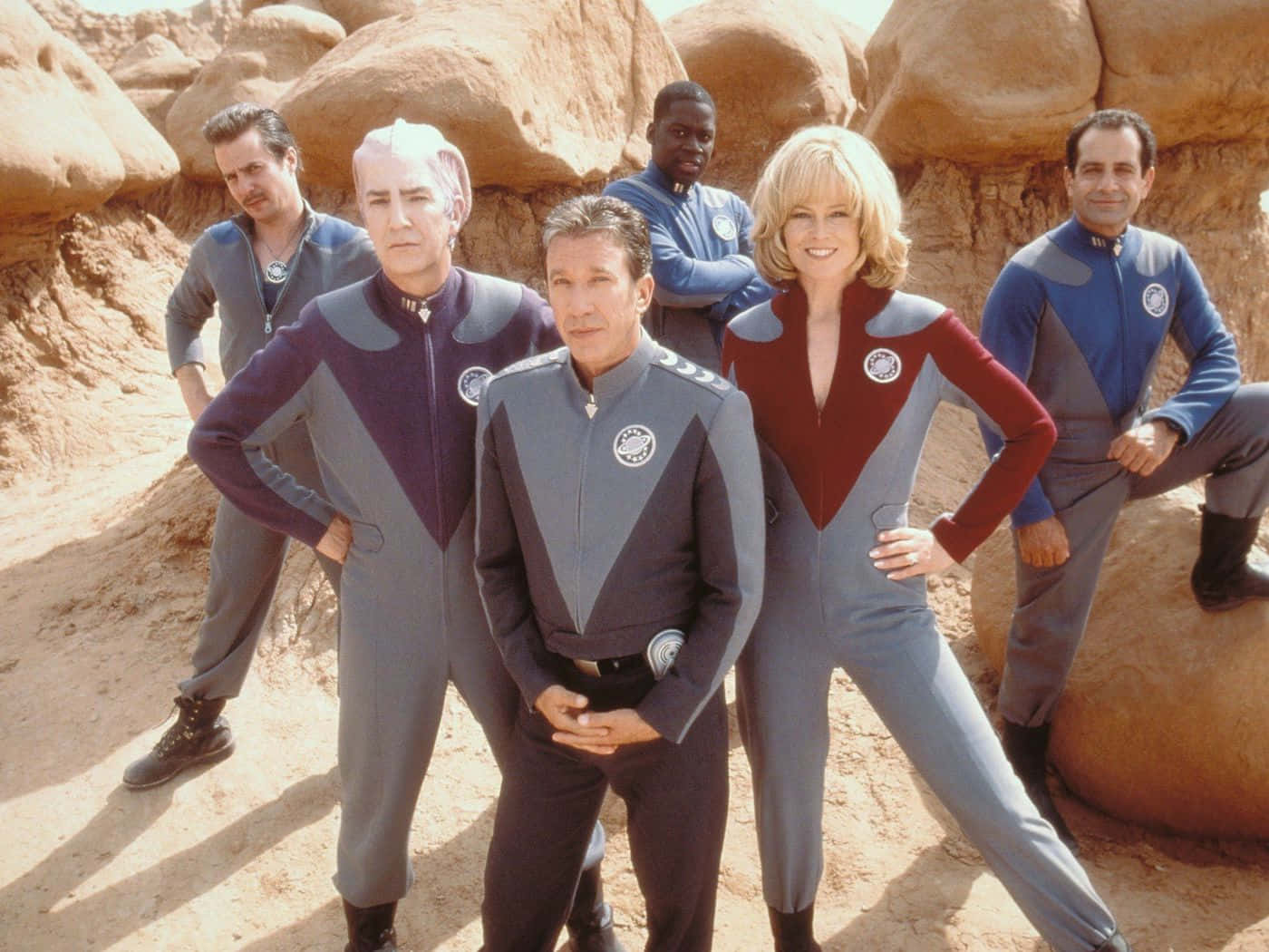 Galaxy Quest 桌布 壁紙 — 六名身穿藍色和灰色制服的工作人員在陽光明媚的沙漠峽谷中自信地擺姿勢，背景是紅色岩層。