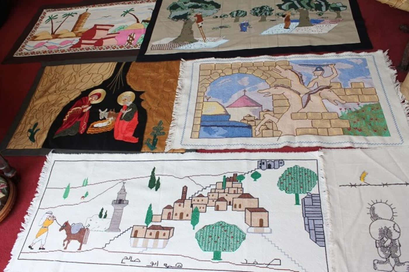 巴勒斯坦 桌布 壁紙 — 六塊刺繡紡織面板描繪聖經和文化場景，色彩鮮豔，邊緣帶流蘇，鋪在紅色表面上。