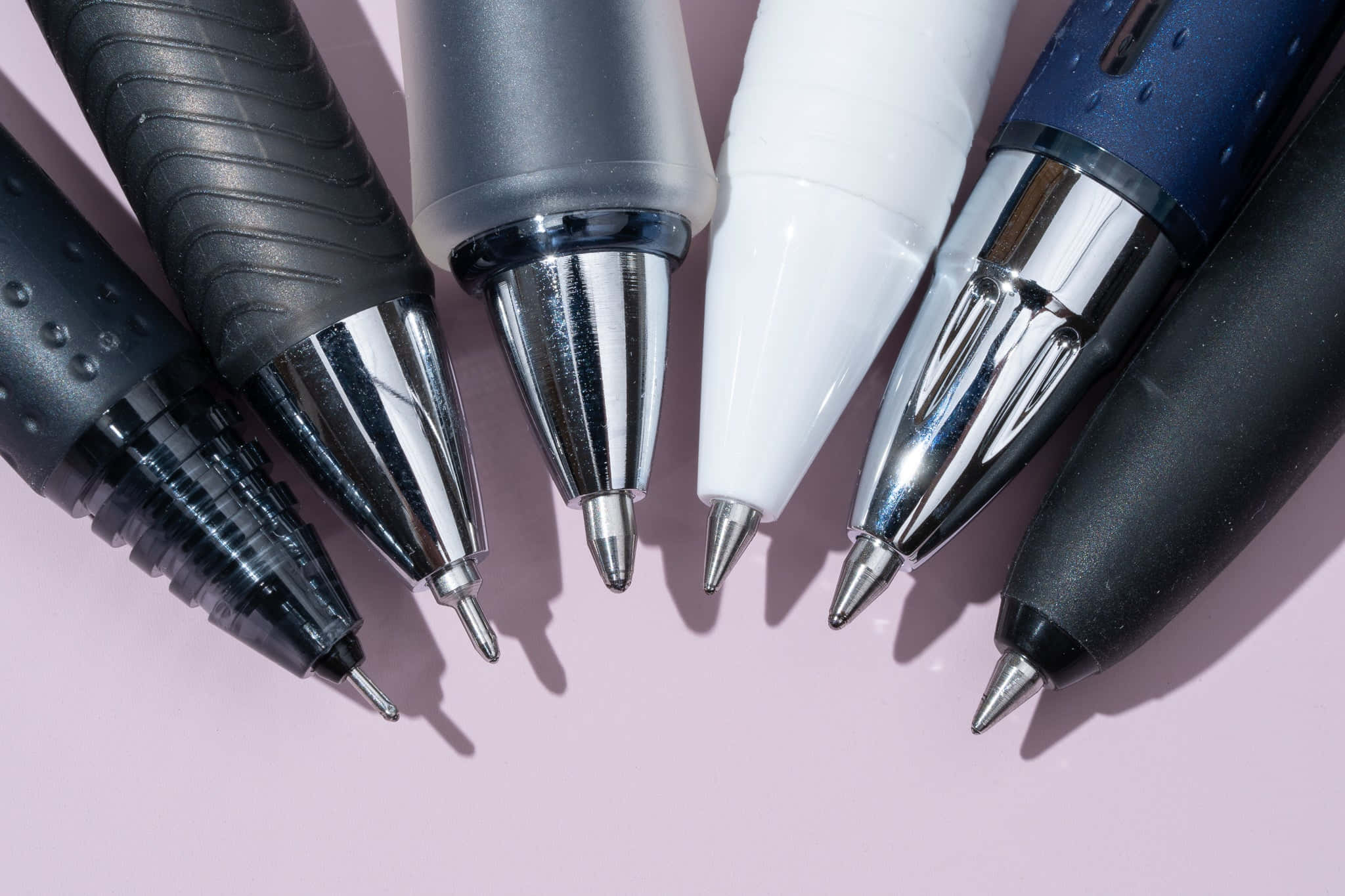 Pens 桌布 壁紙 — 六支帶金屬筆尖的筆以對角線排列在柔軟的粉紅色表面上，投射出清晰的陰影。
