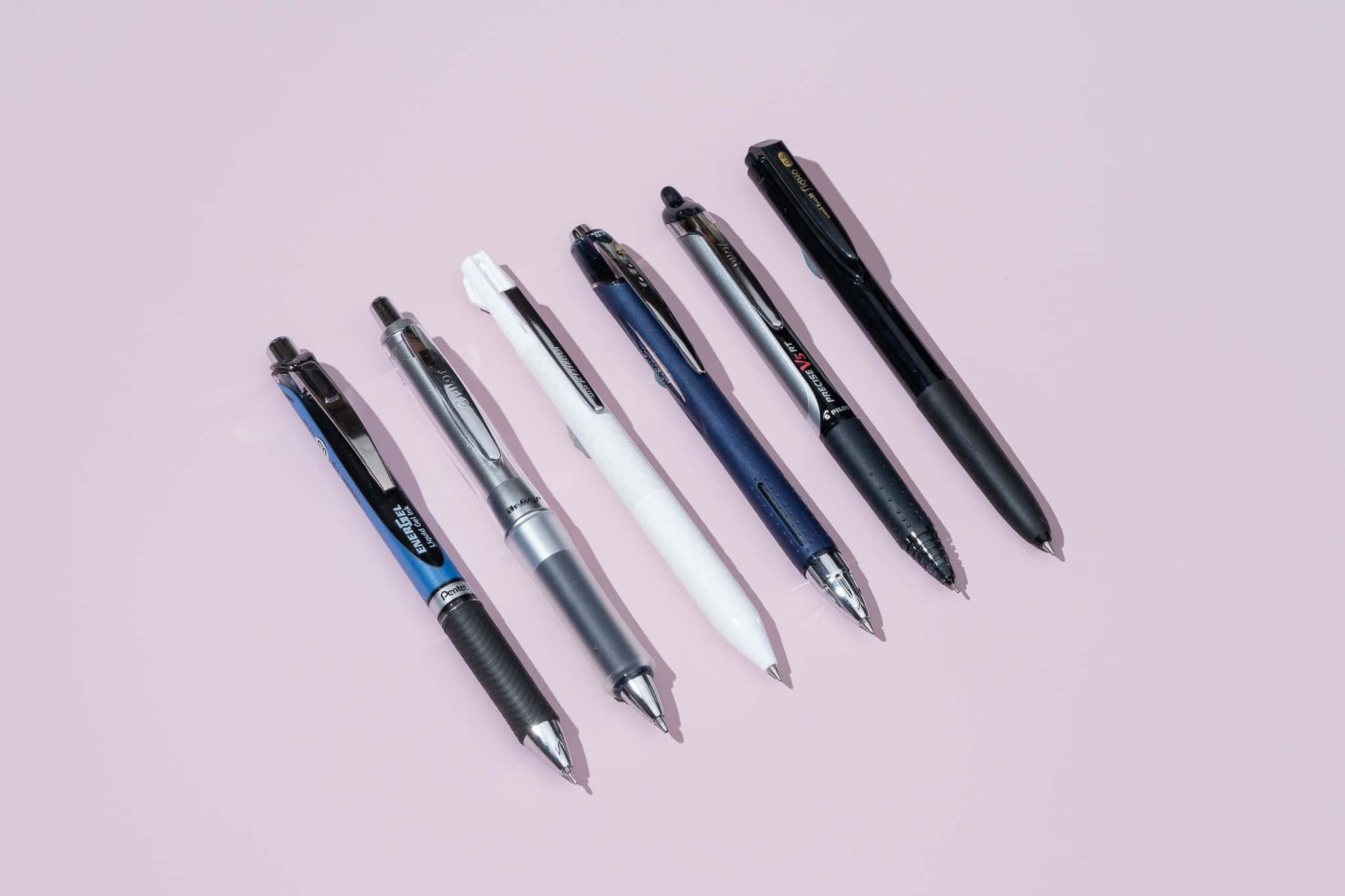Pens 桌布 壁紙 — 六支時尚筆，藍、白、銀、黑色，斜向排列在柔和的粉紅色背景上。