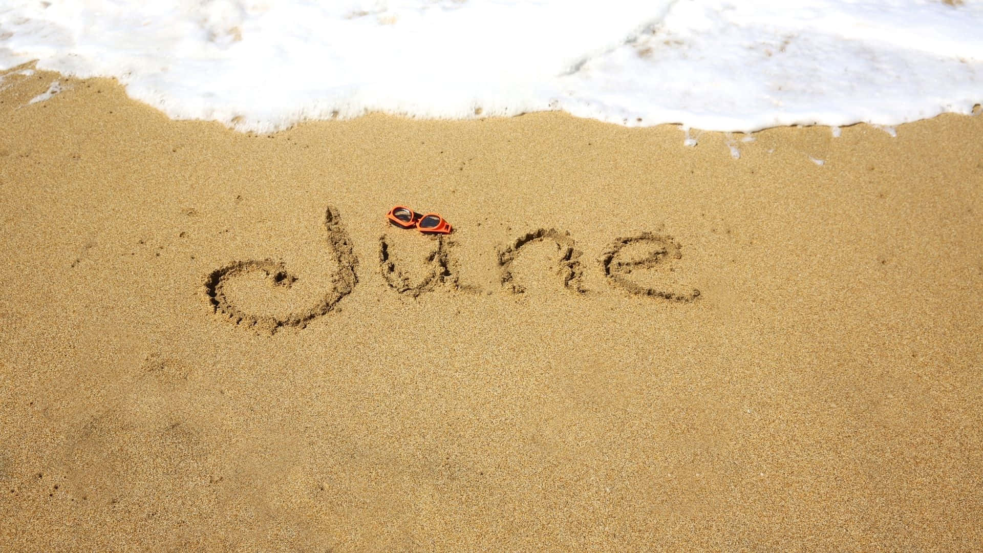 June 桌布 壁紙 — 「六月」這個詞寫在海灘邊緣的濕沙上，一副紅色太陽眼鏡擱在字母U上，溫柔的海浪緩緩湧入。