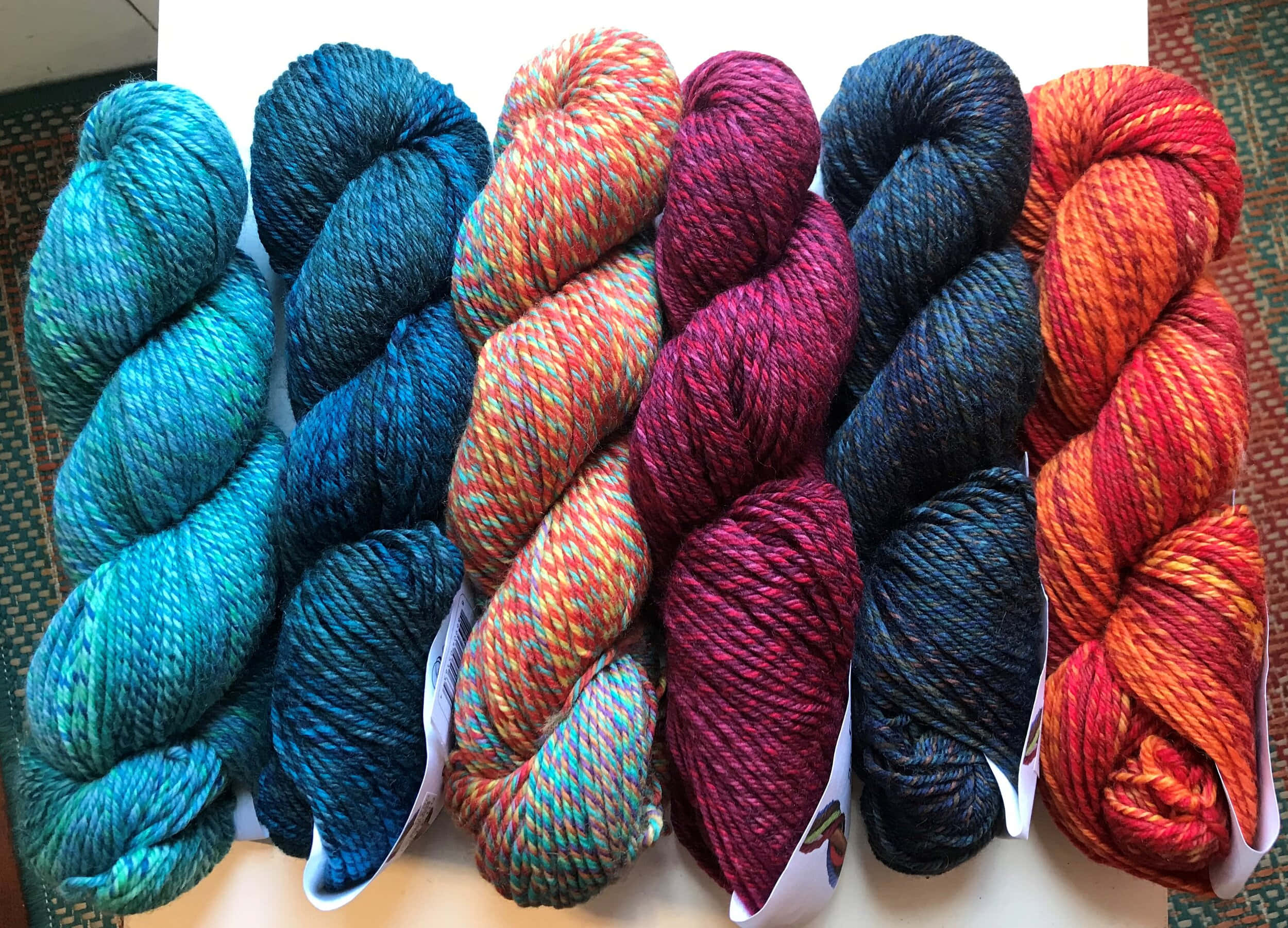 Knitting 桌布 壁紙 — 六束鮮豔的手染紗線，呈現藍綠色、栗紅色和橙色調，整齊地排列在編織墊上。