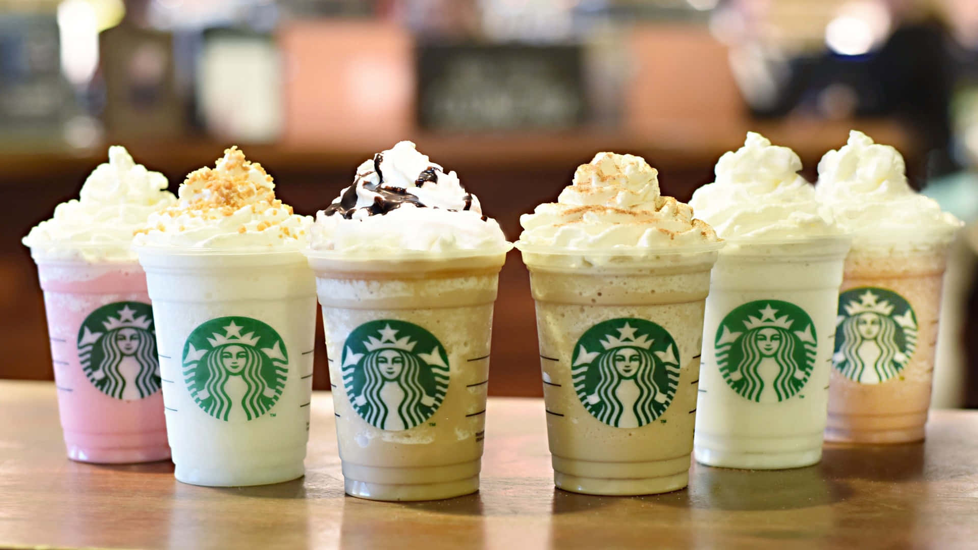 Aesthetic Starbucks 桌布 壁紙 — 六杯星巴克星冰樂，配上鮮奶油和配料，排列在木製櫃檯上。