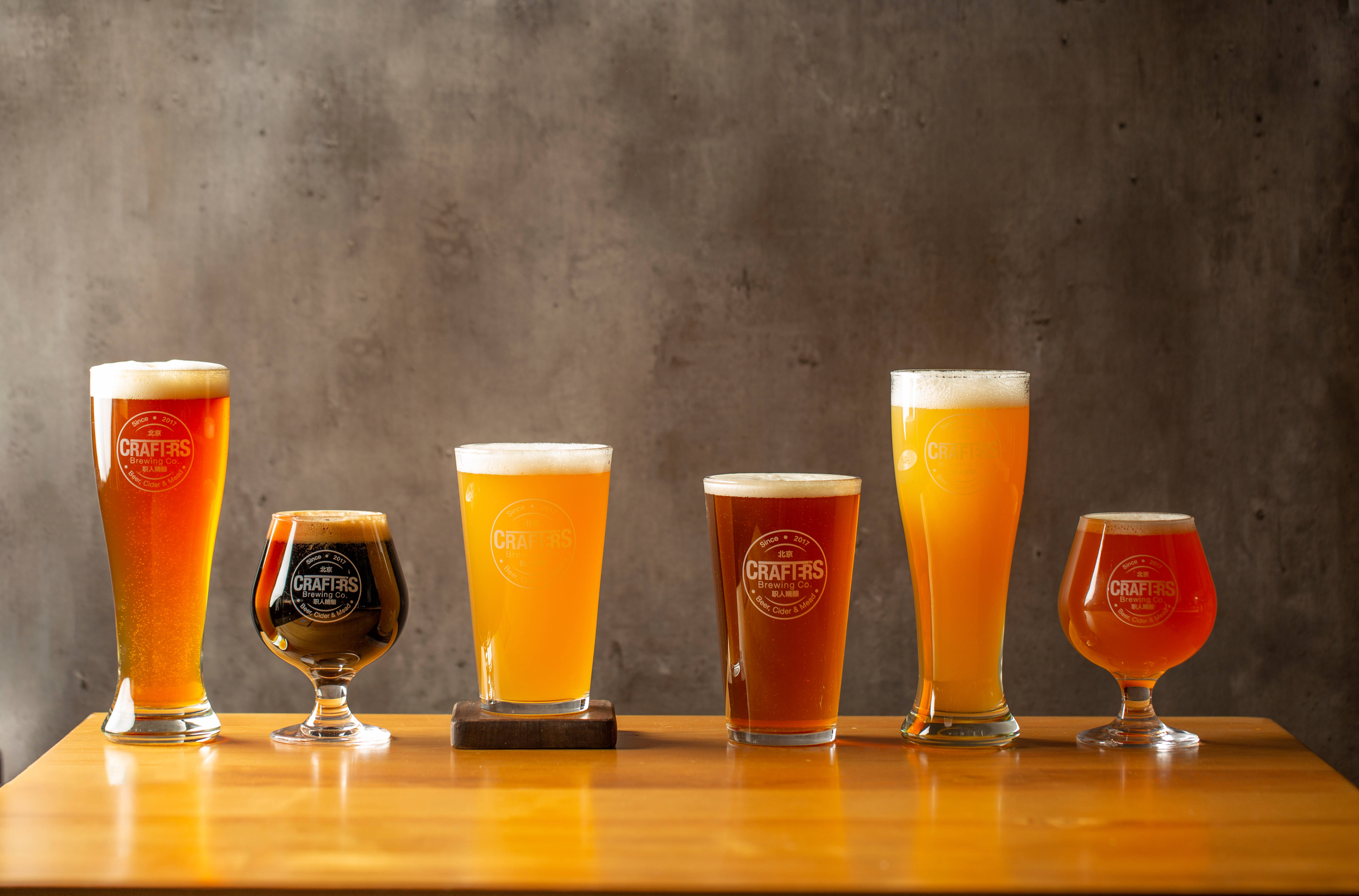 Beer 桌布 壁紙 — 六杯琥珀色和金色的啤酒，帶有泡沫頂部，整齊地放在溫暖的木桌上，背景是紋理灰色牆壁。