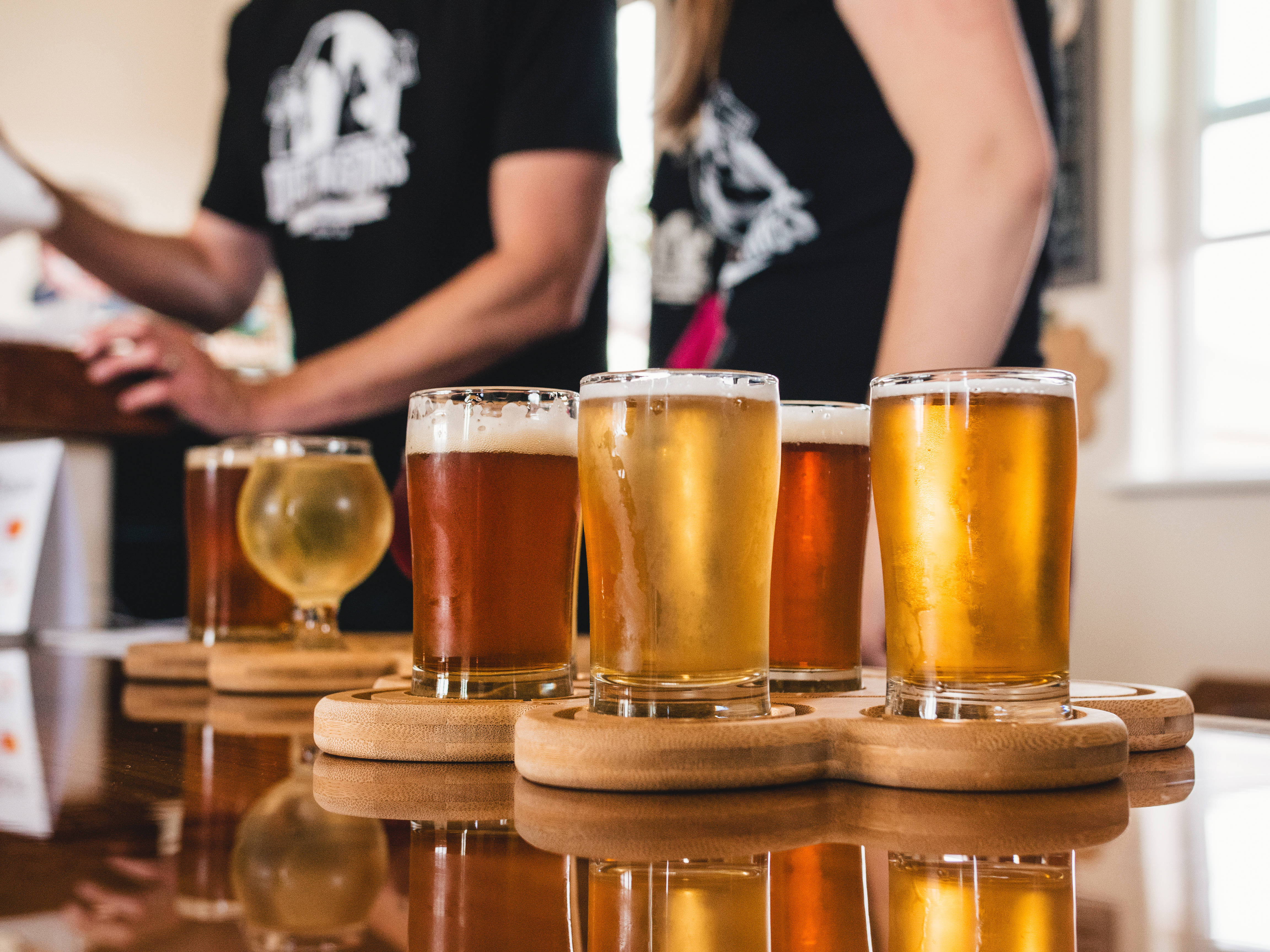 Beer 桌布 壁紙 — 六杯琥珀色和金色的啤酒放在木杯墊上，在拋光的吧檯上反射出倒影，背景中身穿黑色襯衫的調酒師模糊不清的特寫。