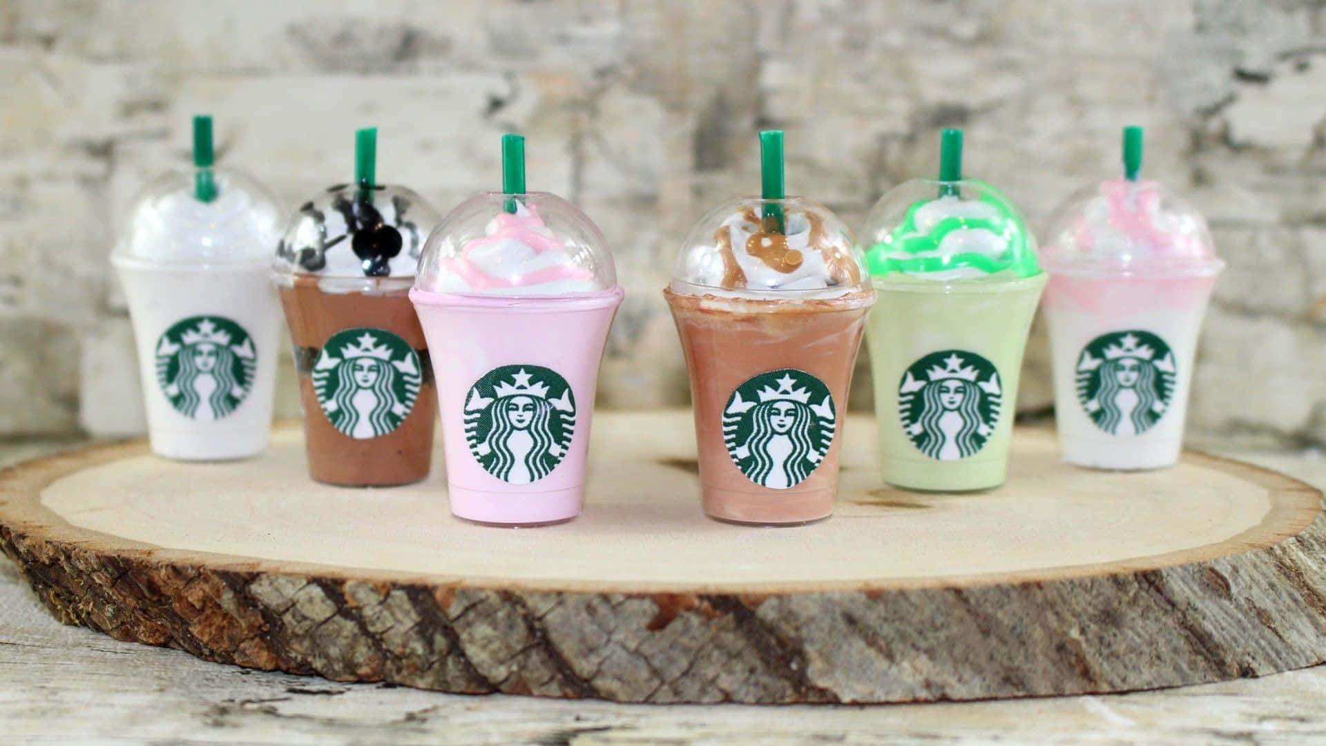 Aesthetic Starbucks 桌布 壁紙 — 六杯色彩繽紛的星巴克星冰樂，配上鮮奶油和綠色吸管，整齊排列在一塊質樸的木板上，背景是風化的磚牆。