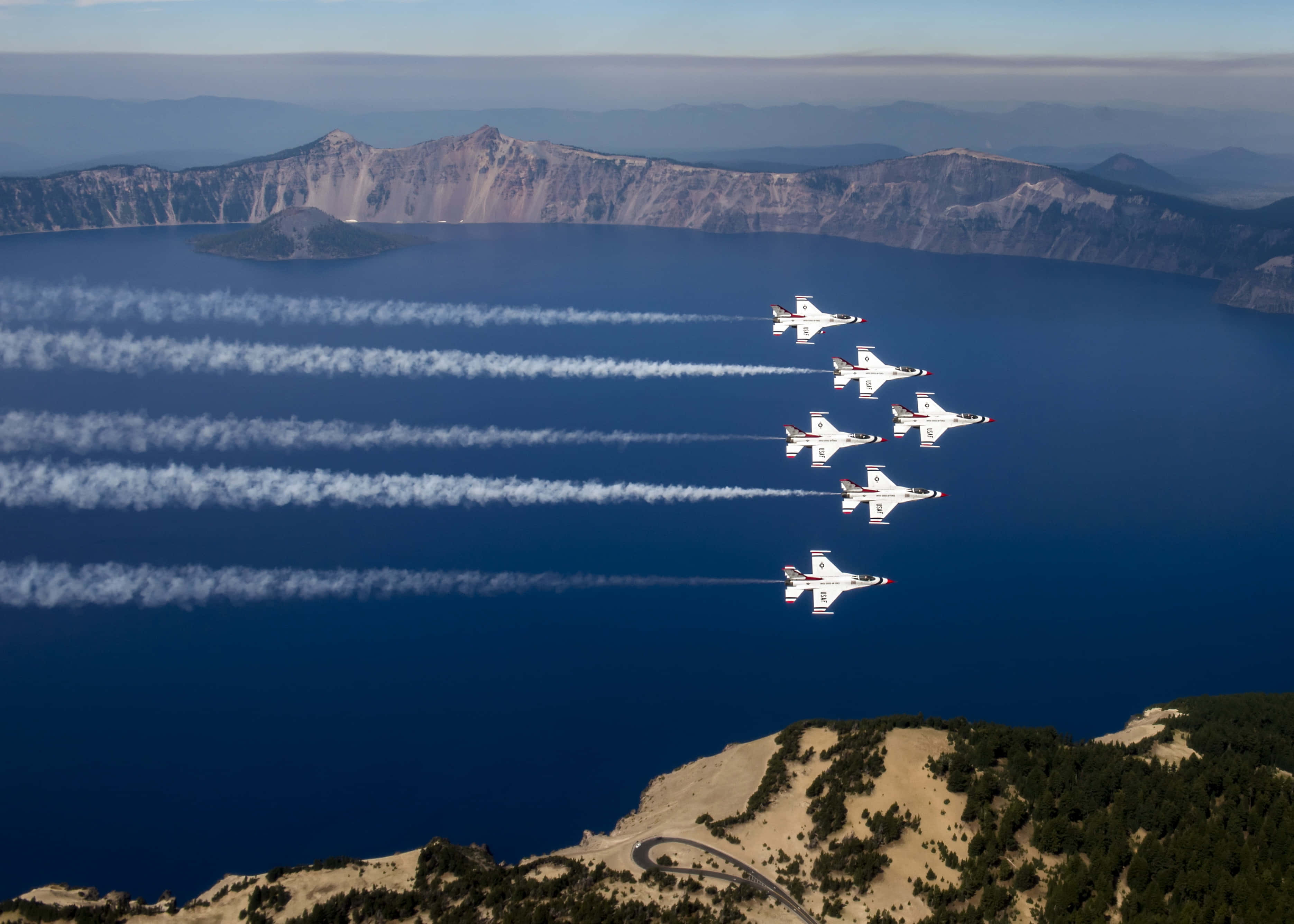 Air Force 桌布 壁紙 — 六架白色噴射機在火山口湖上空緊密編隊飛行，在深藍色水域和崎嶇火山邊緣留下白色煙霧痕跡。
