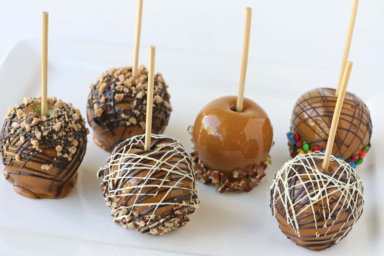 Caramel Apples 桌布 壁紙 — 六款美食焦糖蘋果，搭配堅果、巧克力醬和彩色糖果等各式配料，整齊排列在白色盤子上。