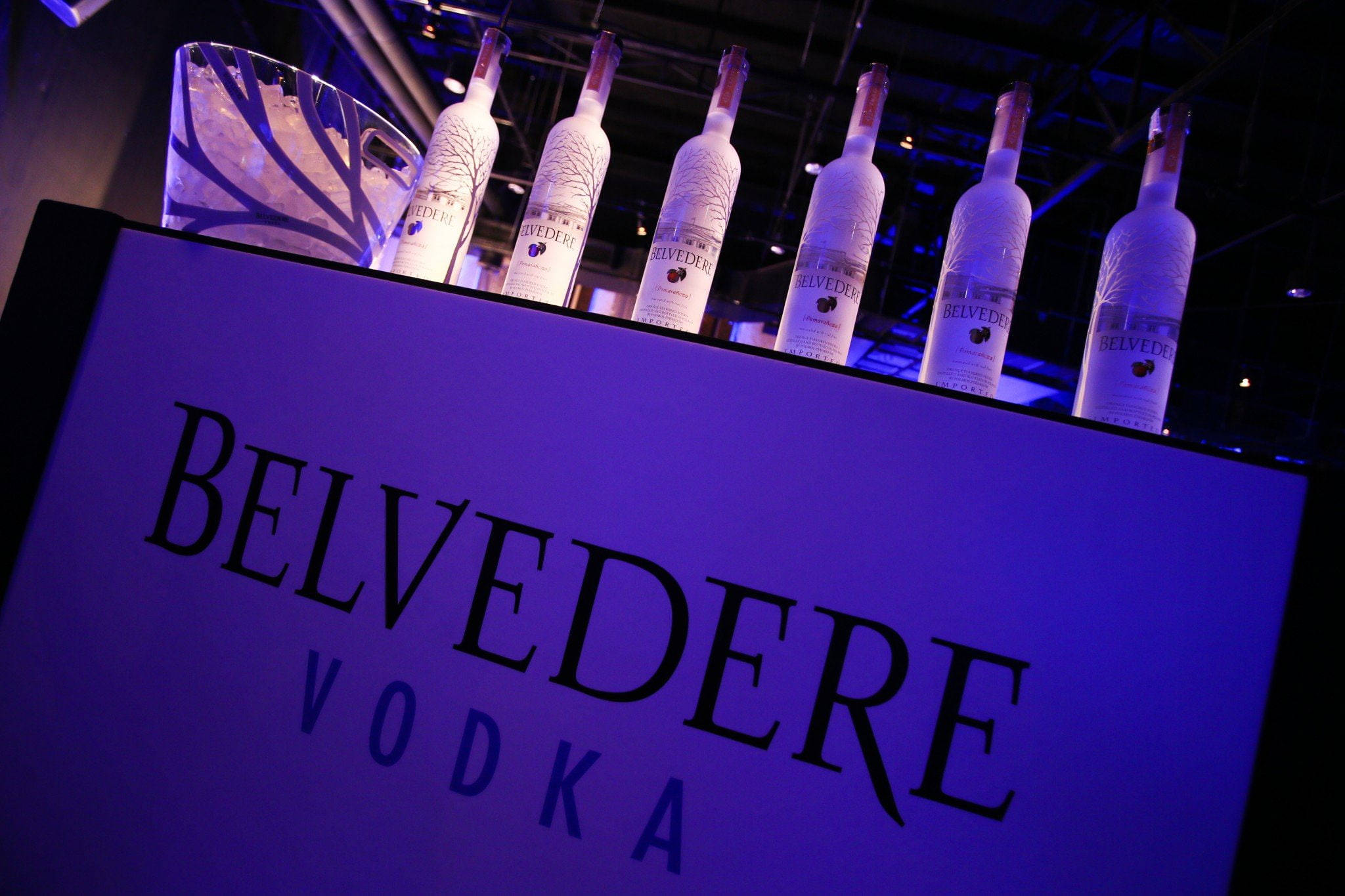alcohol 桌布 壁紙 — 六瓶Belvedere伏特加酒在紫色燈光下閃閃發光，放在品牌展示架上，旁邊有一個冰桶。