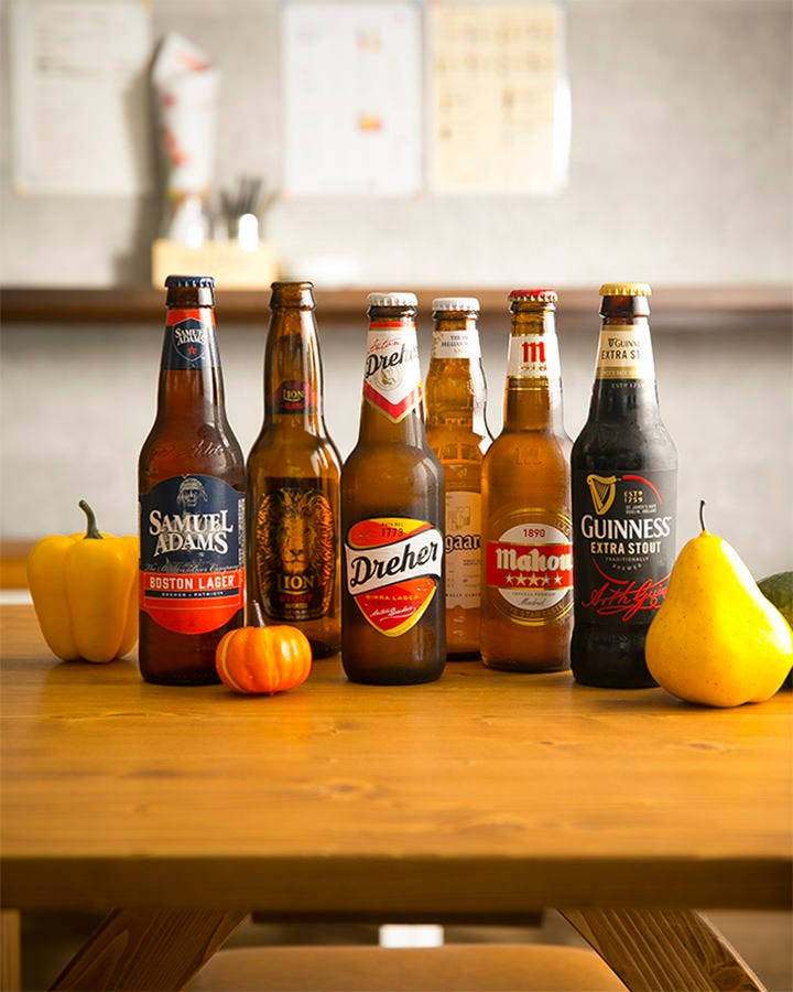 alcohol 桌布 壁紙 — 六瓶啤酒和秋季水果整齊地擺放在一張木桌上，處於溫暖照明的廚房環境中。