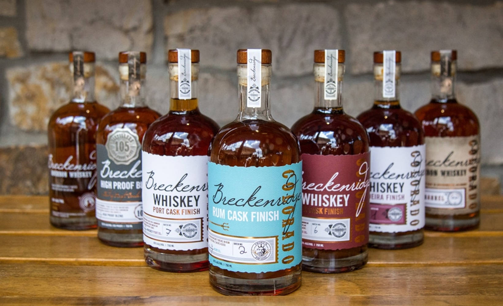 drinks 桌布 壁紙 — 六瓶帶有彩色標籤的Breckenridge威士忌排成一排，放在木質表面上，背景是石牆。