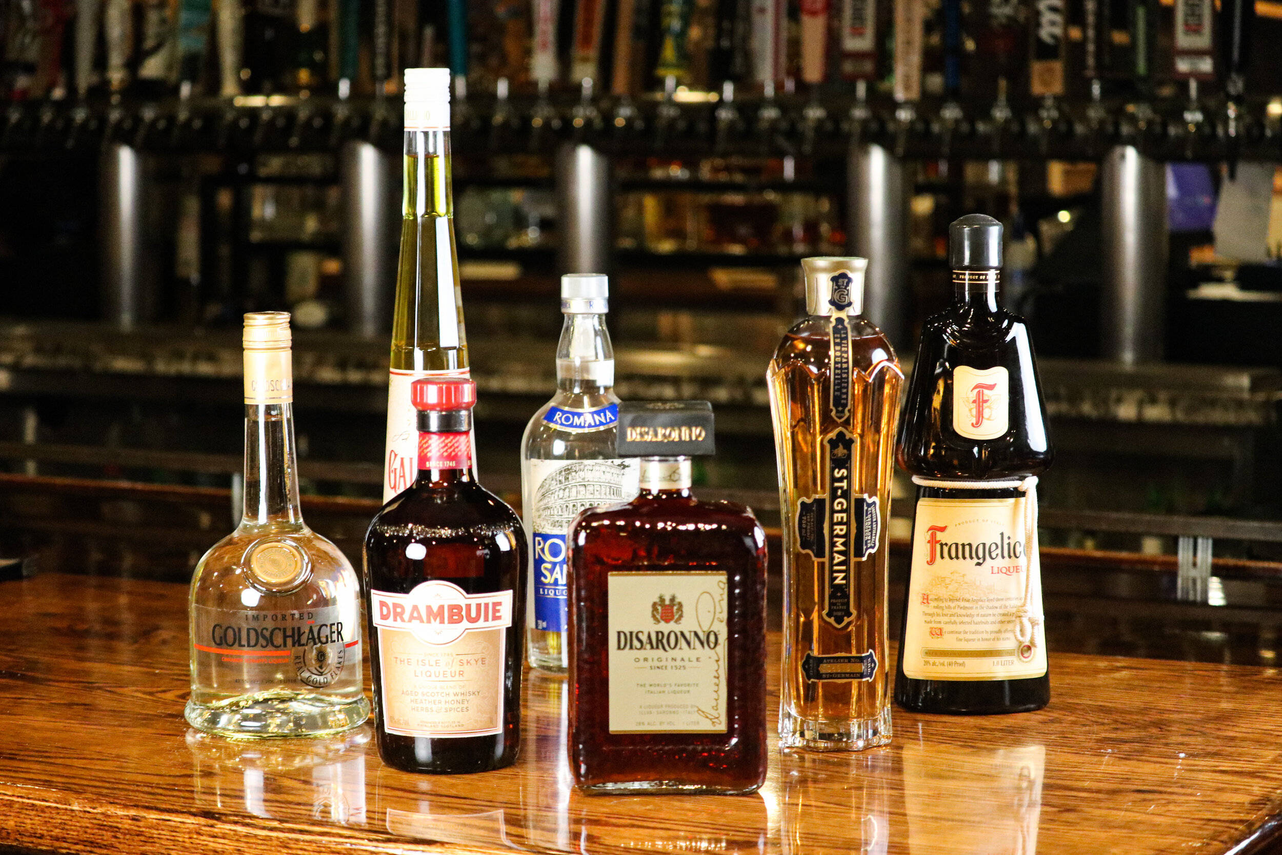 alcohol 桌布 壁紙 — 六種不同的酒瓶整齊地排列在拋光木製吧檯上，背景是模糊的吧檯龍頭和架子。