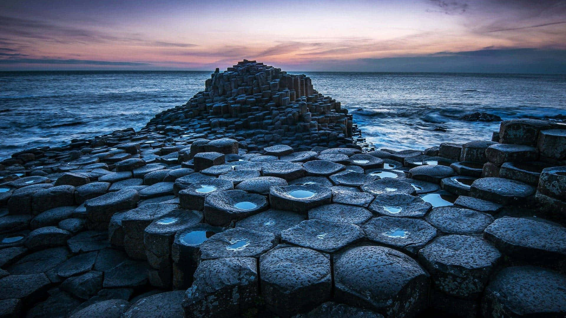 Rocks 桌布 壁紙 — 六角形玄武岩柱在黃昏時從海洋中升起，上方是紫色和藍色的天空。