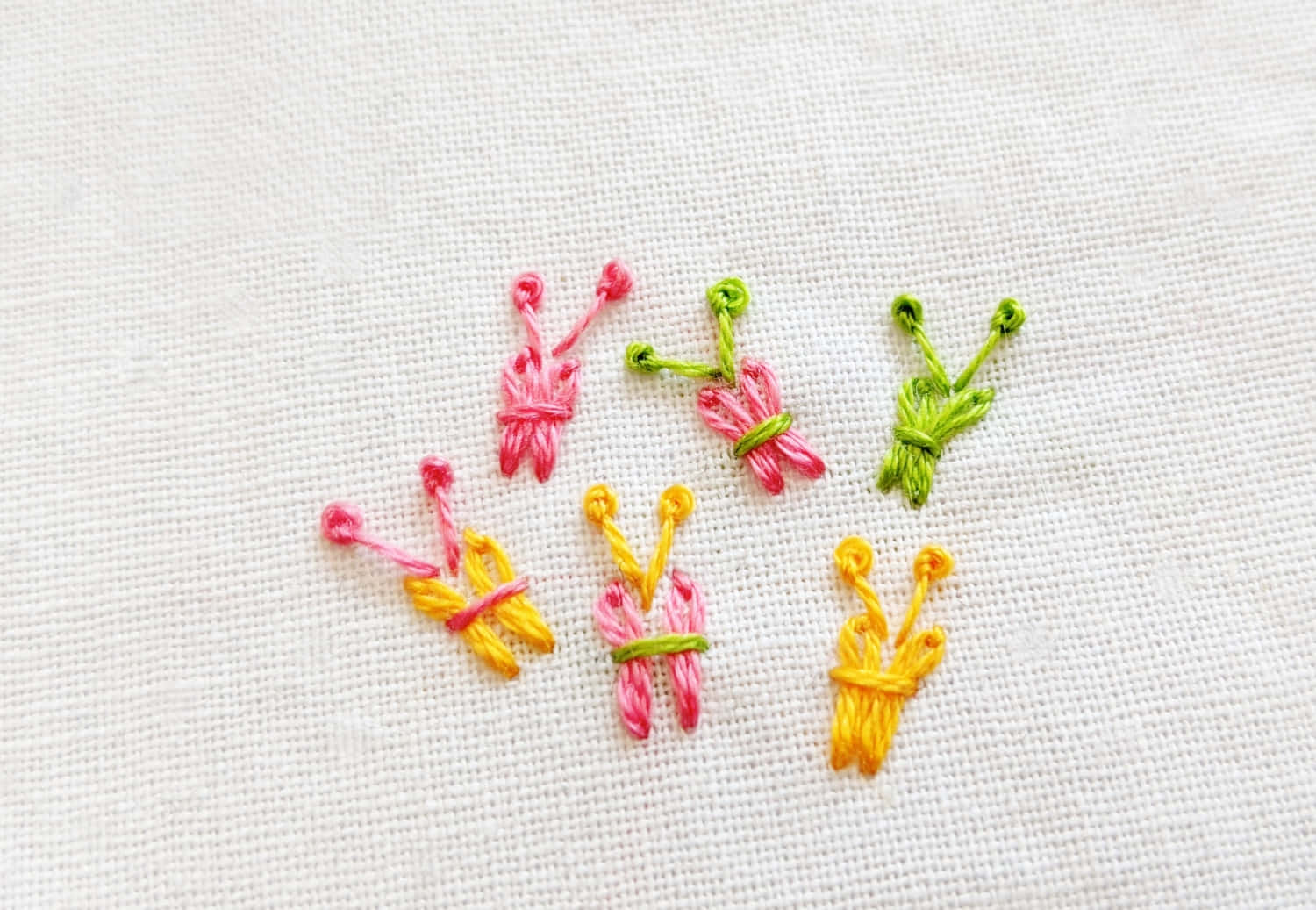 Butterfly Stitch 桌布 壁紙 — 六隻粉紅色、黃色和綠色的細小彩色刺繡蝴蝶散落在有紋理的白色布料上。