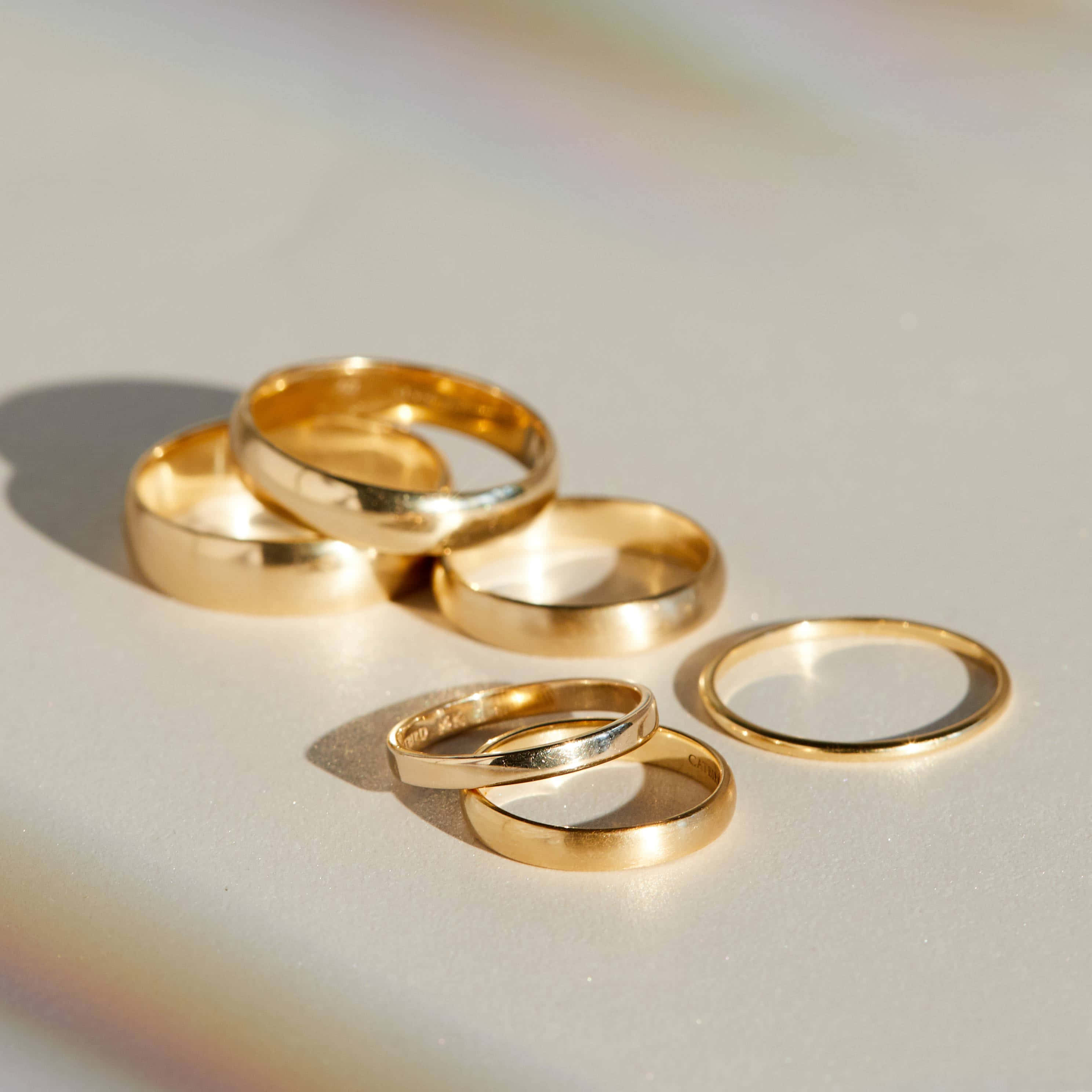 Wedding Rings 桌布 壁紙 — 六隻金色結婚戒指在柔和陽光下，於淺色表面上閃耀，帶有柔和陰影和彩虹光折射。