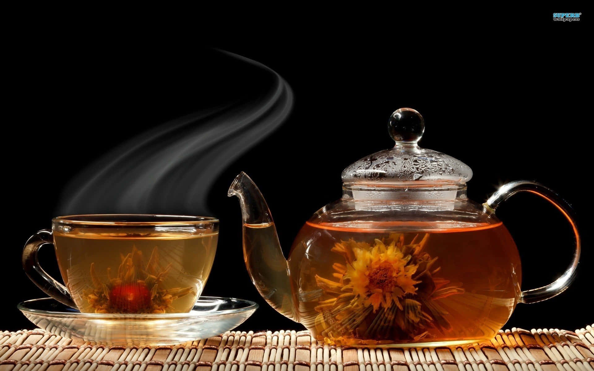 tea 桌布 壁紙 — 冒煙的玻璃茶壺和杯子，裝滿金色花草茶和盛開的花朵，放在黑色背景前的編織墊上。