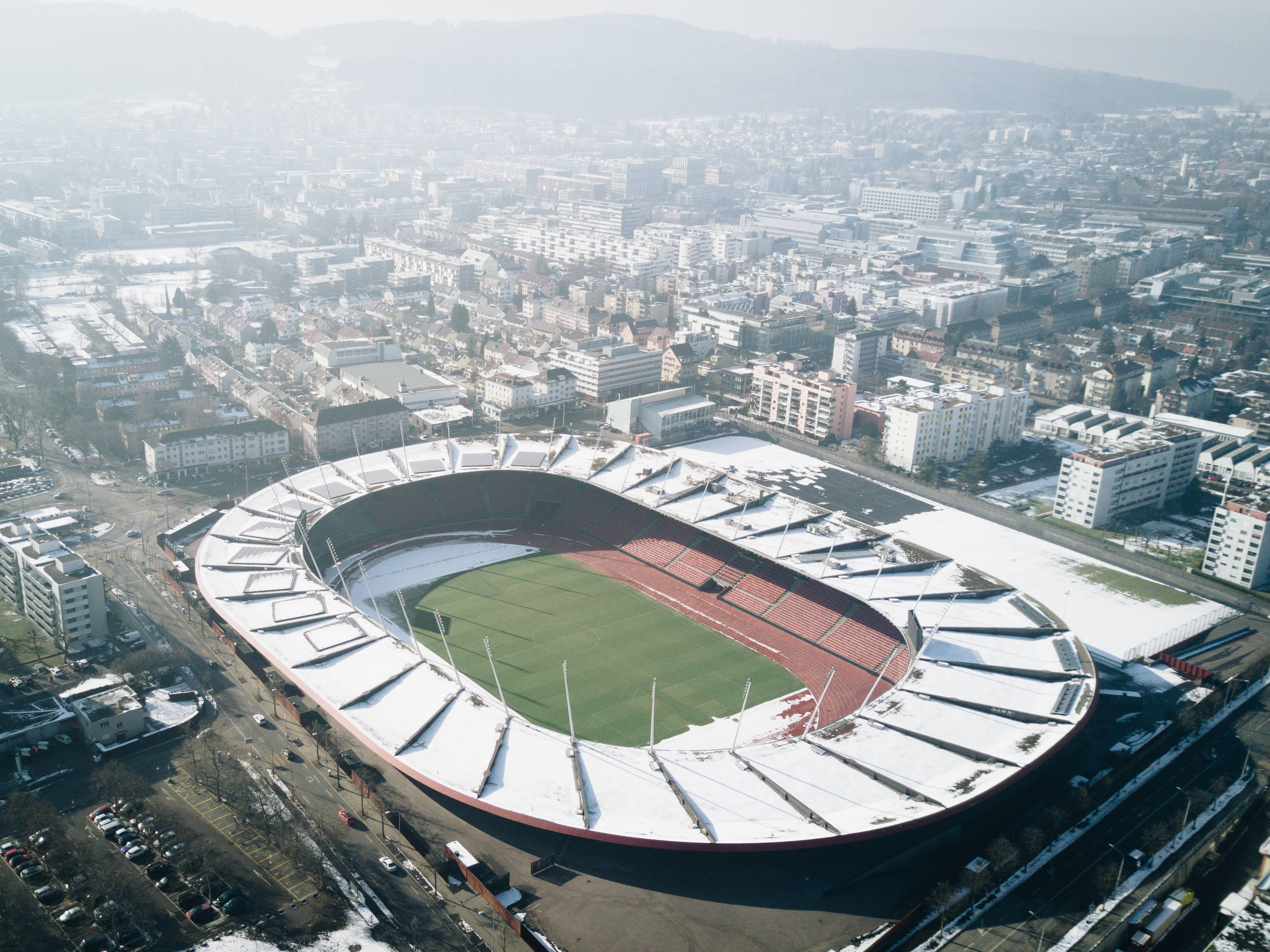 Olympics 桌布 壁紙 — 冬季城市景觀中，一個被雪覆蓋的體育場，內有綠色場地和紅色跑道，鳥瞰圖。