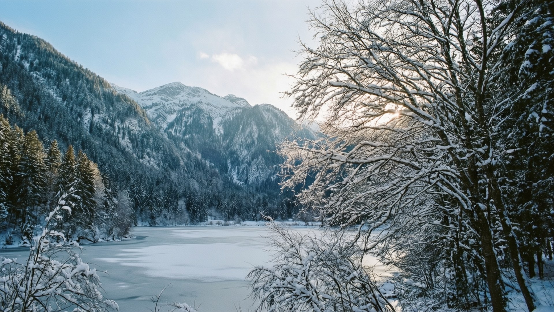 冬日仙境 壁紙 — 一個結冰的湖泊坐落在白雪覆蓋的山脈中，光禿禿的樹木在清澈的藍天下框出寧靜的冬日景色。