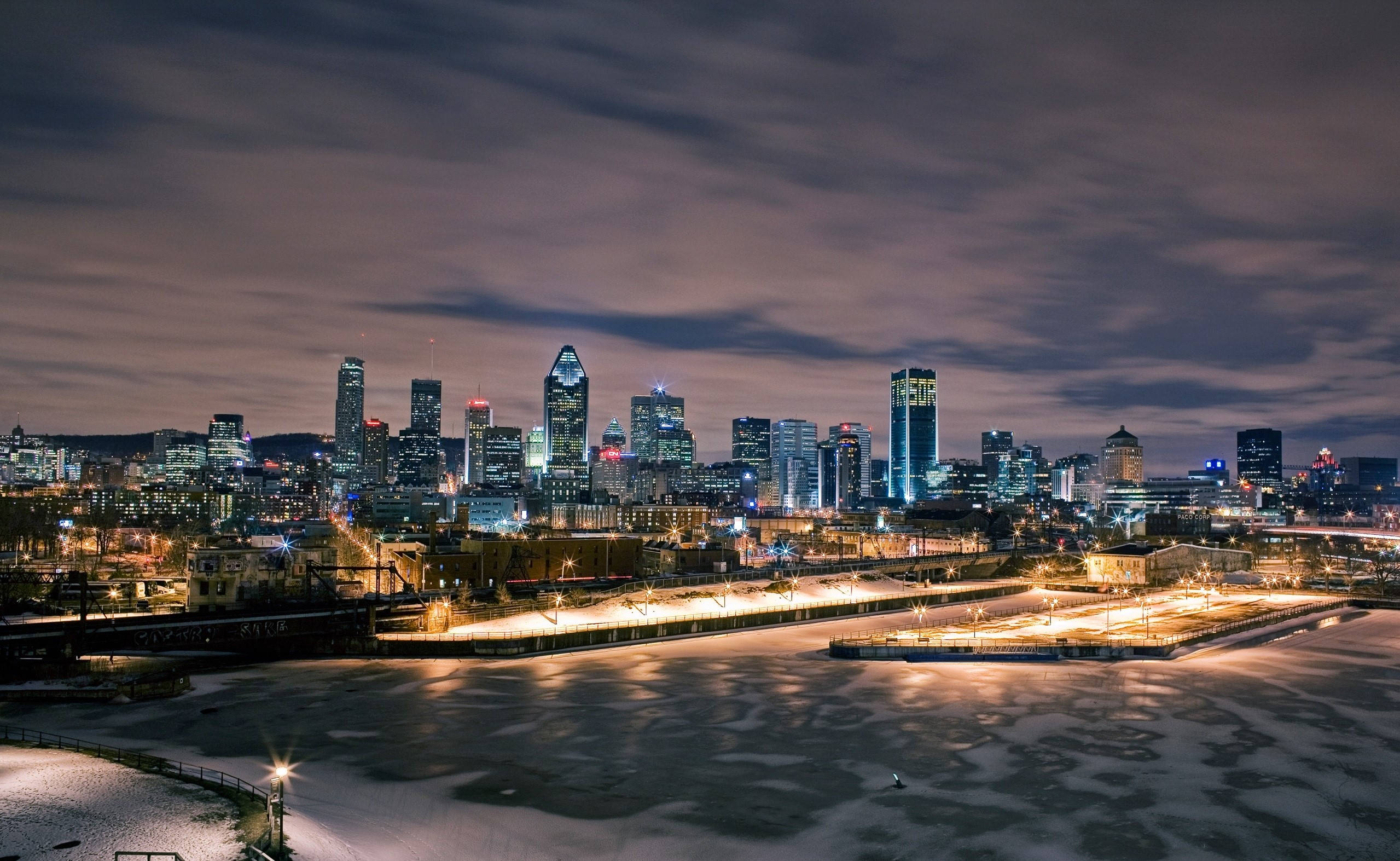 Montreal 桌布 壁紙 — 冬日夜晚的城市景觀，雪覆蓋的地面和冰冷的河流反射著高聳摩天大樓的霓虹燈光，天空多雲。