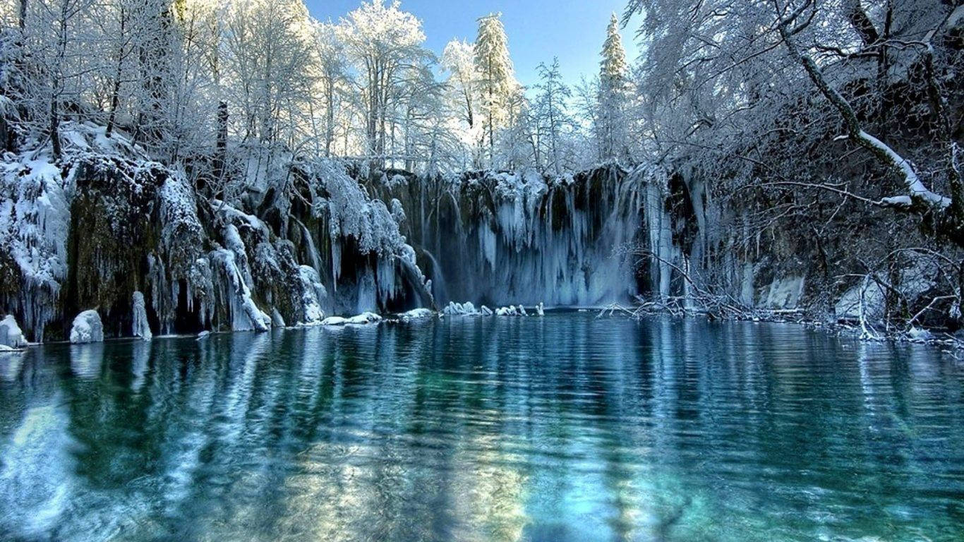 Snow Desktop 桌布 壁紙 — 冰凍瀑布流入一個清澈的綠松石色水池，周圍是白雪覆蓋的樹木，在明亮的藍天下。