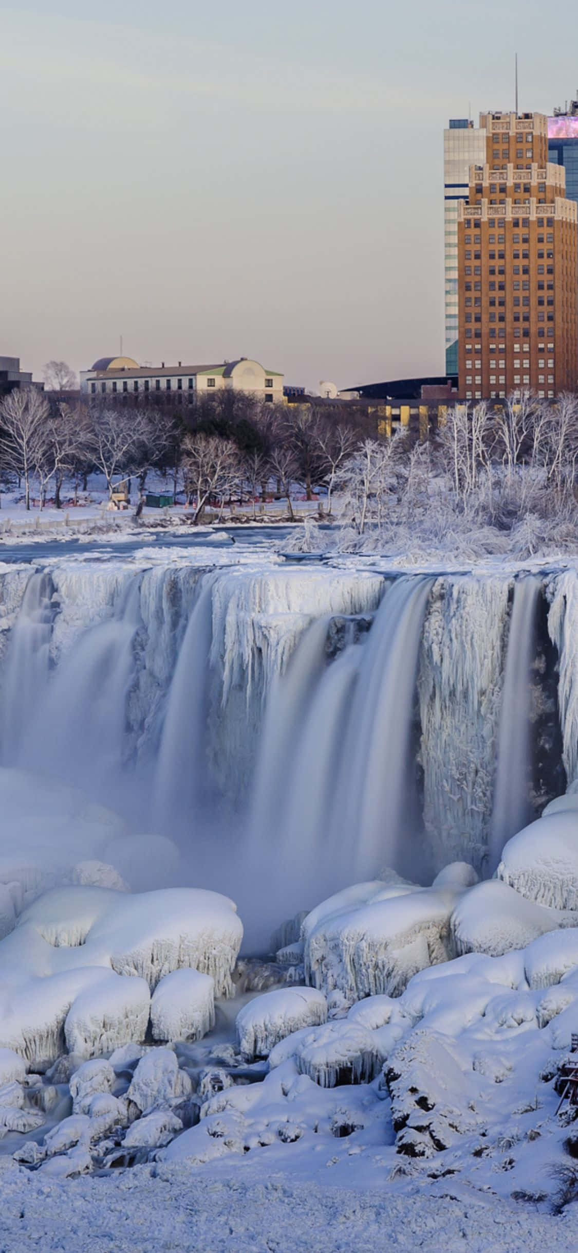Niagara Falls 桌布 壁紙 — 冰凍瀑布流過被雪覆蓋的岩石，背景有冰層結構和城市建築，在黎明時分。