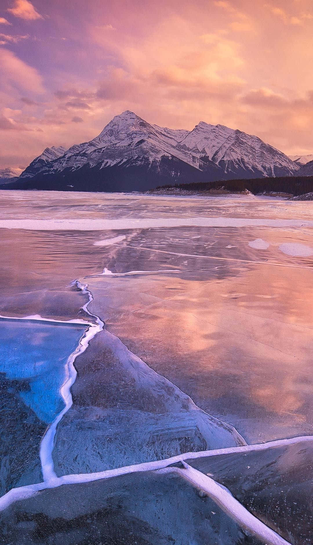 Frozen Lake 桌布 壁紙 — 冰凍的湖面反射出鮮豔的粉紅色和橙色日落，白雪覆蓋的山脈在背景中聳立。