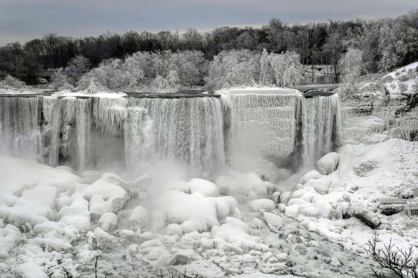 720p Niagara Falls 桌布 壁紙 — 冰凍的瀑布在灰色的冬日天空下，流過冰冷的岩石和積雪的樹木。