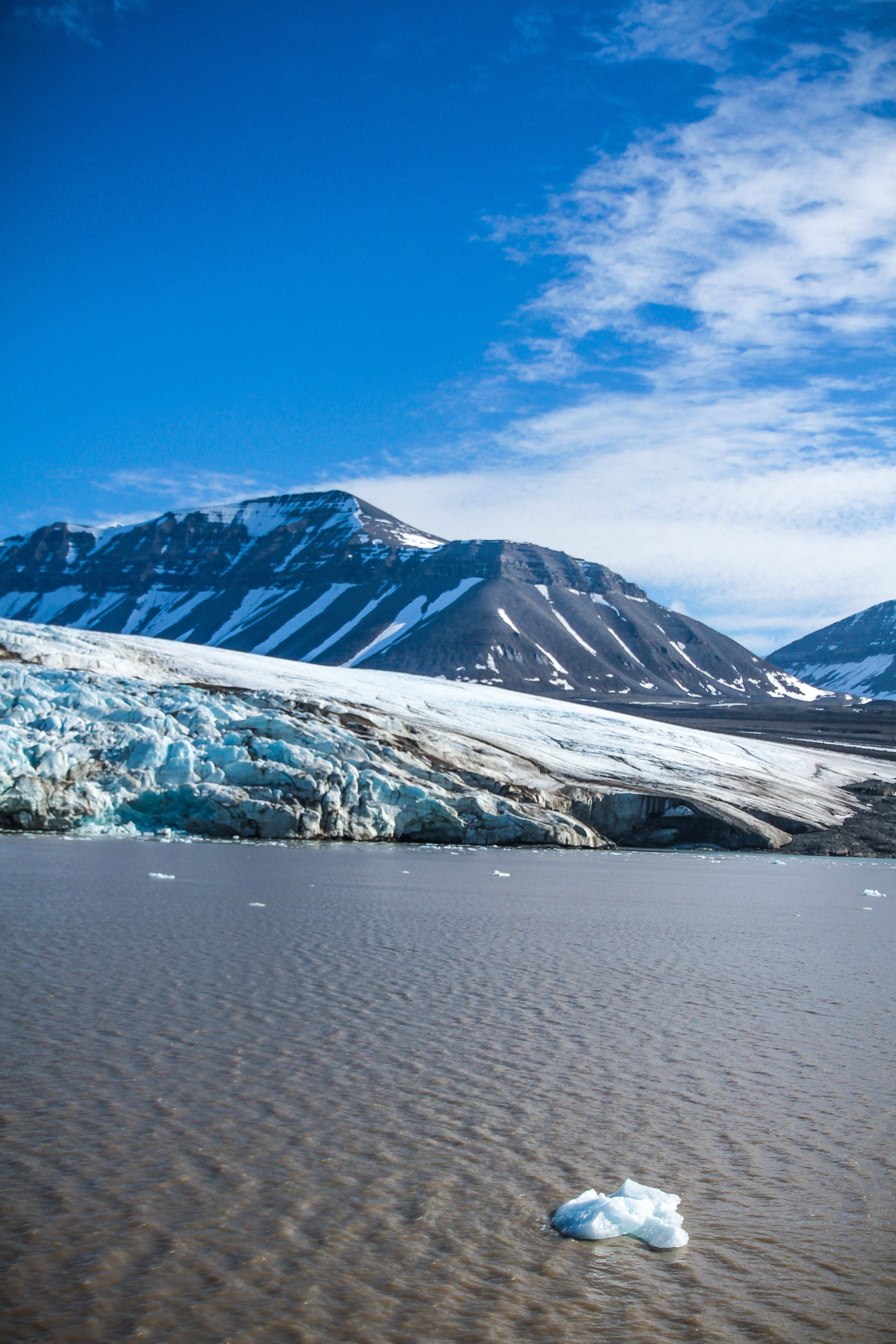Svalbard 桌布 壁紙 — 冰川在明亮的藍天下崩解到平靜的棕色湖泊中，背景是積雪覆蓋的山脈。