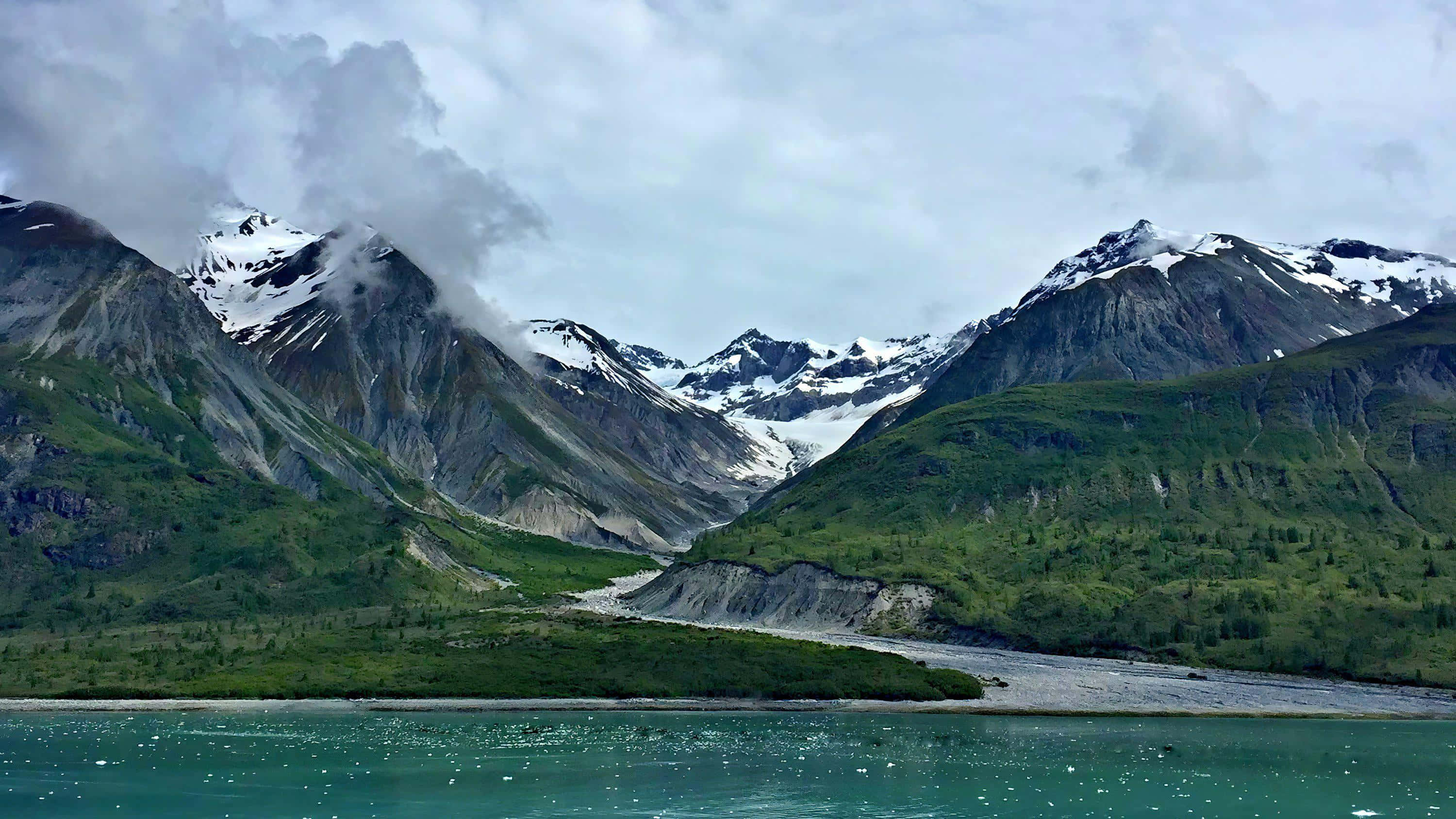 山景 桌布 壁紙 — 冰川雕刻的山谷，積雪覆蓋的山峰聳立在綠松石色的河流之上，天空多雲。