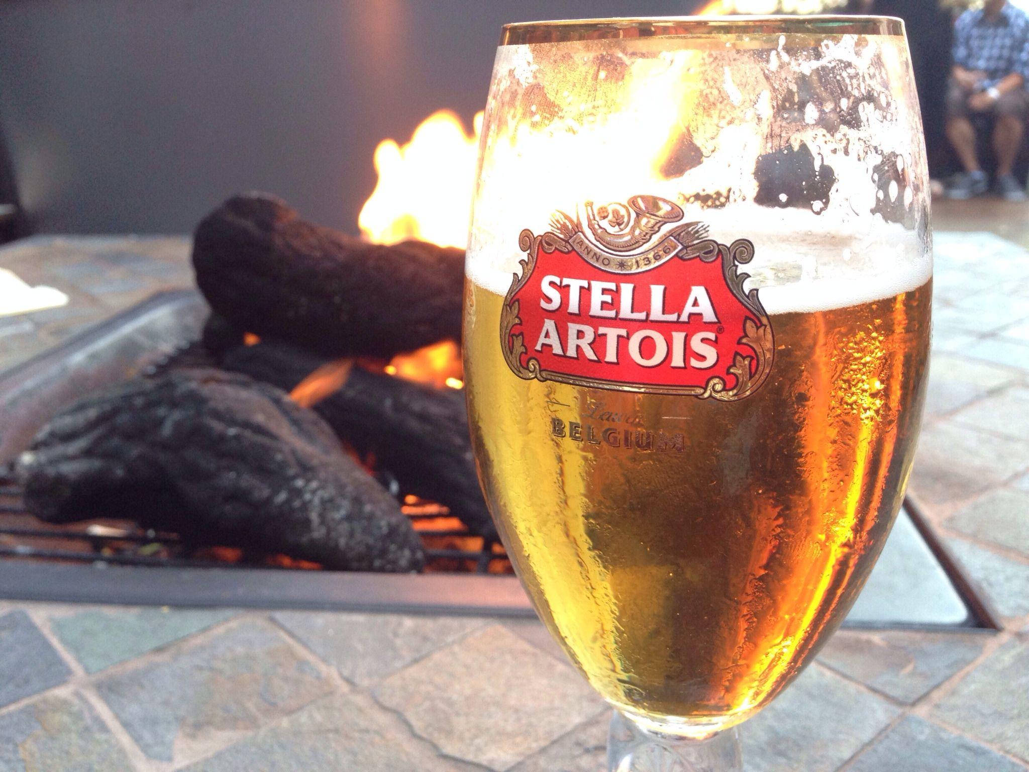 Beer 桌布 壁紙 — 冰涼的 Stella Artois 啤酒在石頭露台上閃爍著冷凝水珠，旁邊是噼啪作響的戶外火坑。