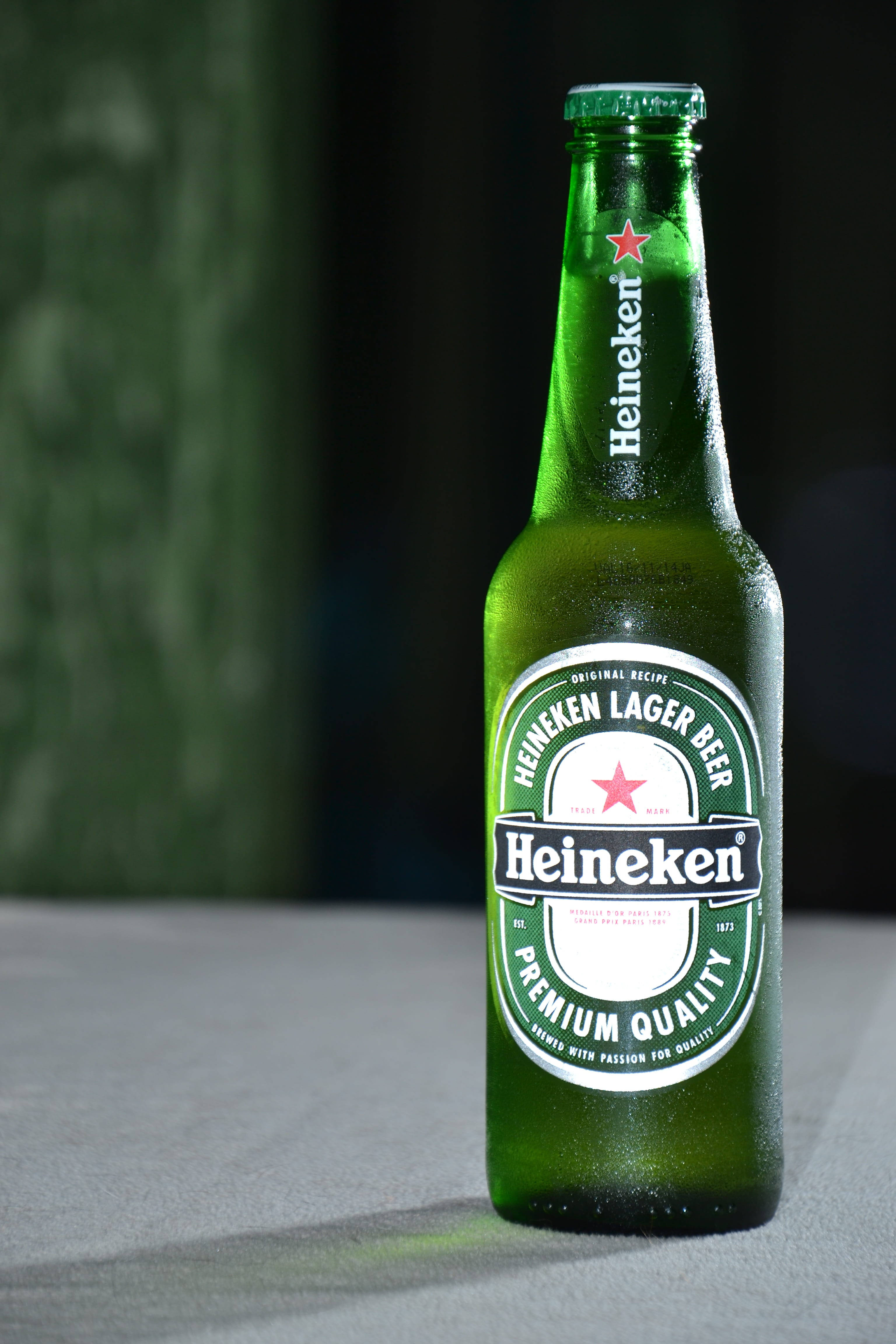 Drink 桌布 壁紙 — 冰涼的海尼根啤酒瓶上凝結的水珠在柔和燈光下閃爍，背景為深綠色紋理。