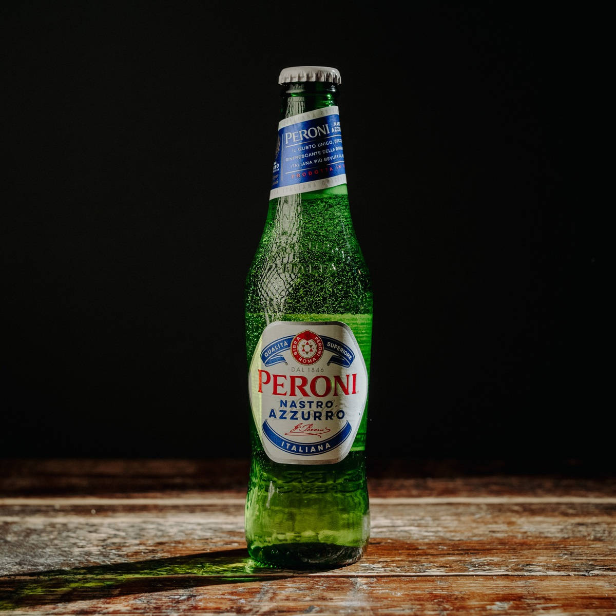 Beer 桌布 壁紙 — 冰涼的綠色 Peroni Nastro Azzurro 啤酒瓶在深色背景下的質樸木質表面上閃爍著冷凝水珠。
