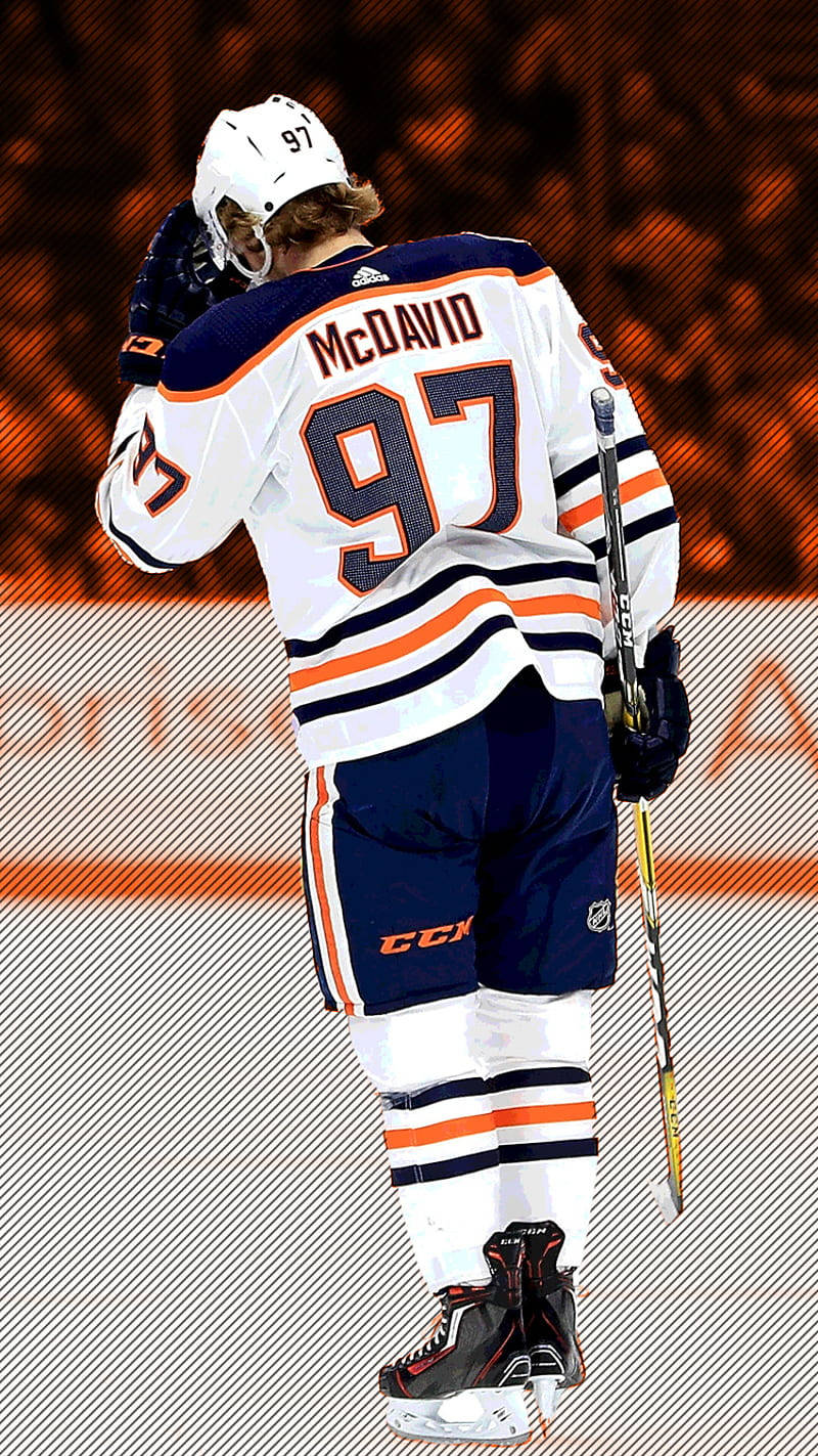 hockey 桌布 壁紙 — 冰球運動員 Connor McDavid 身穿白色、橙色和藍色 97 號球衣，站在冰上手持球棍，背景人群模糊。