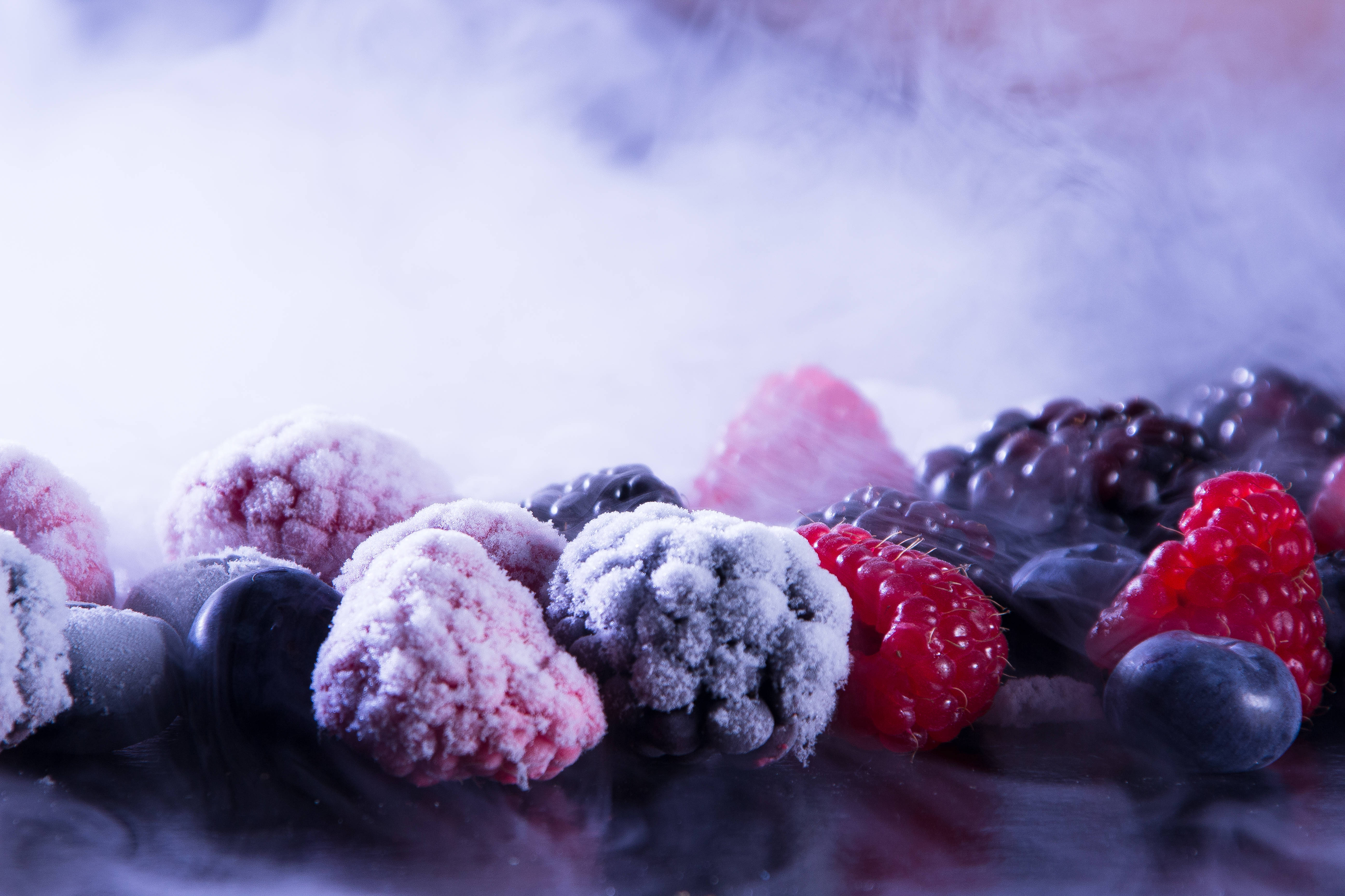 Fruits 桌布 壁紙 — 冷凍莓果覆蓋著霜，在冷紫色燈光下，於黑暗反光表面上的旋轉薄霧中發光。