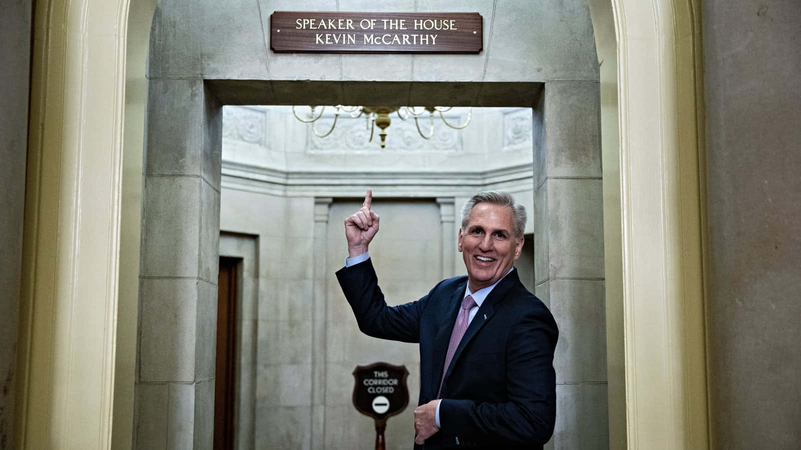 人物 桌布 壁紙 — 凱文·麥卡錫在正式的政府走廊中，穿過一個刻有「Speaker of the House Kevin McCarthy」的巨大石拱門，微笑並向上指。